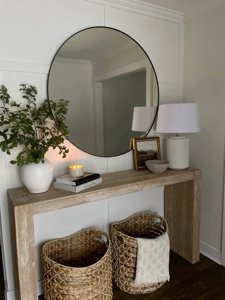 A light-washed wood console table styled with a large black round wall mirror, a white vase with greenery, a lit candle on stacked books, and two tall woven seagrass storage baskets sitting on a dark wood floor.