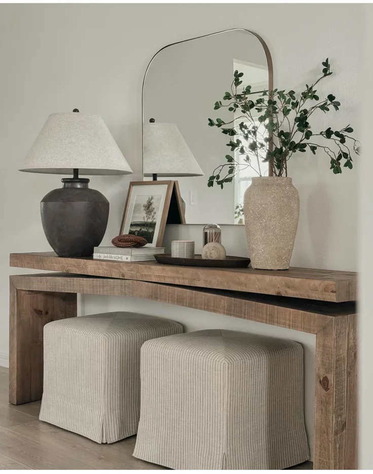 A modern wooden entry table styled with a silver arched wall mirror, a large dark grey ceramic table lamp, a stone vase with green branches, and two striped upholstered footstools underneath.