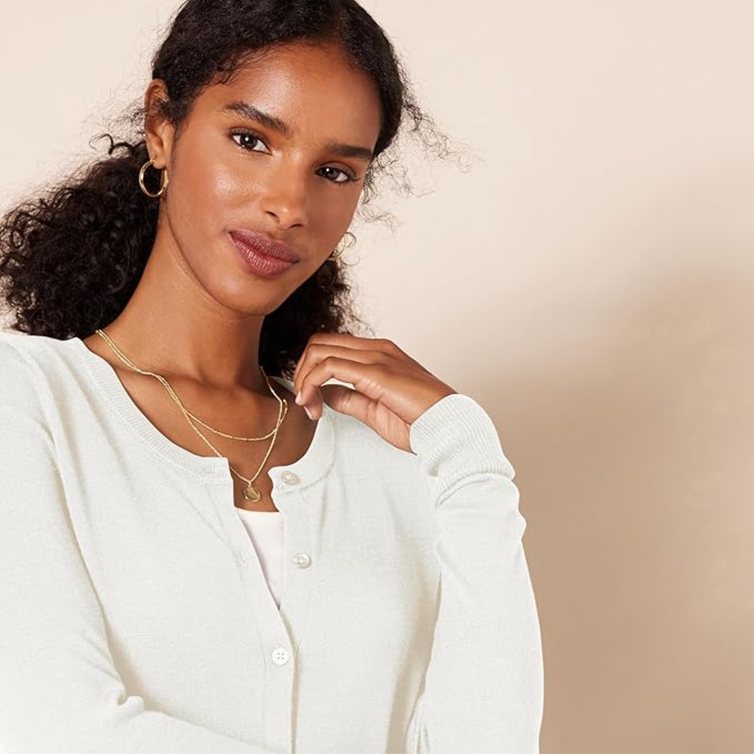 A close-up shot of a woman with curly hair wearing a white knit cardigan, a simple gold coin necklace, and small hoop earrings, looking at the camera against a neutral background.