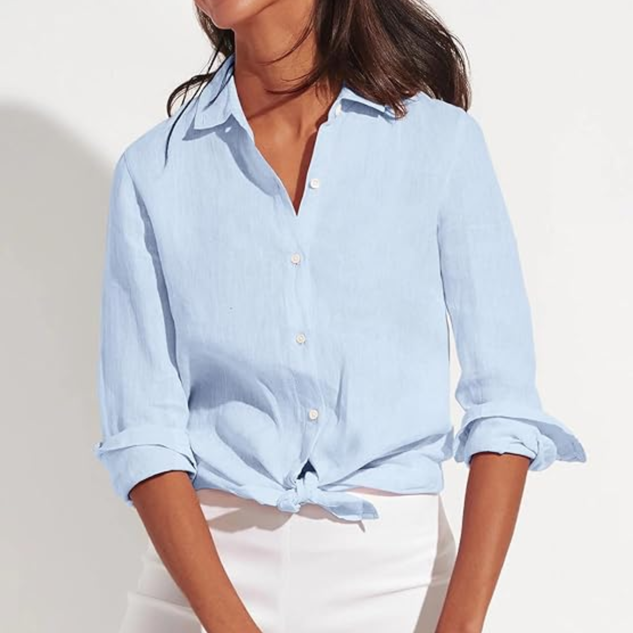 A close-up of a woman's torso wearing a light blue, long-sleeve linen-blend button-down shirt that is tied at the waist, paired with white pants.