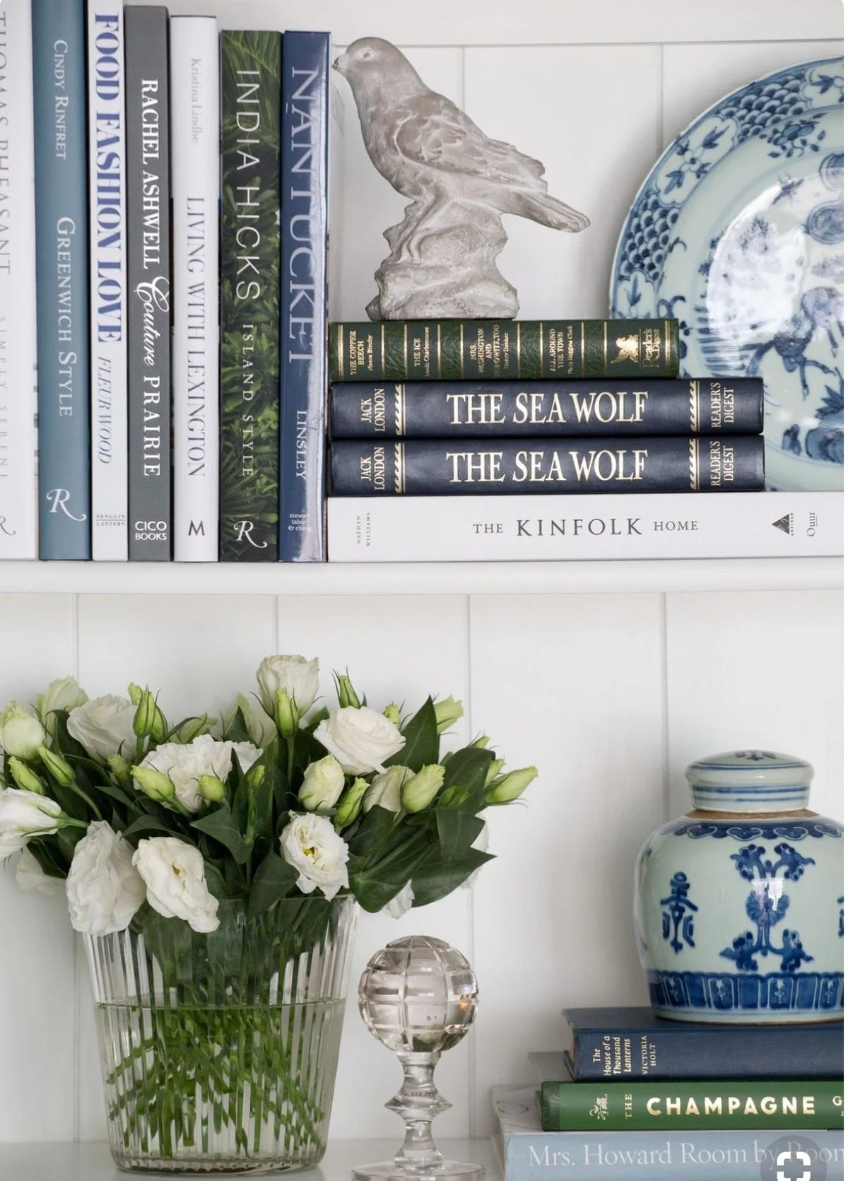 Chinoiserie-inspired home decor display on white shelves featuring blue and white porcelain plates, a ginger jar, a ceramic bird sculpture, the 'Kinfolk Home' book, and fresh white flowers in a ribbed glass vase.