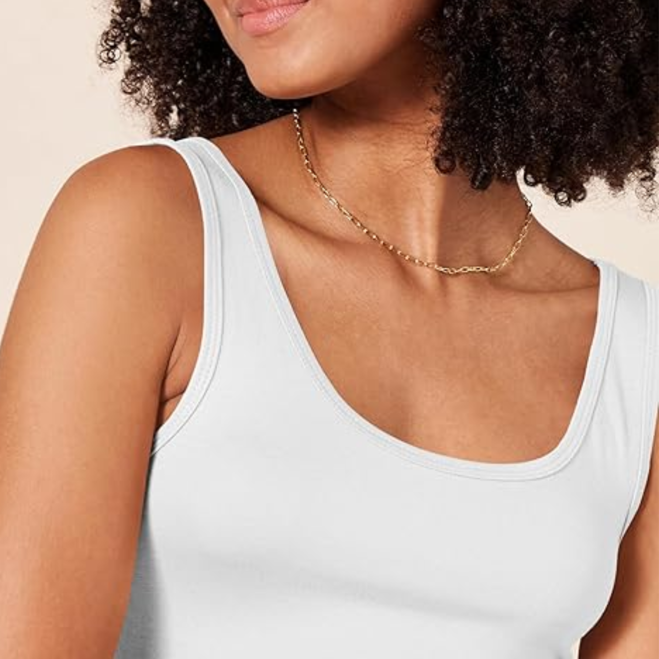 A close-up of a woman's shoulder and neck area, wearing a simple white scoop neck tank top and a delicate gold chain necklace, with dark curly hair visible.