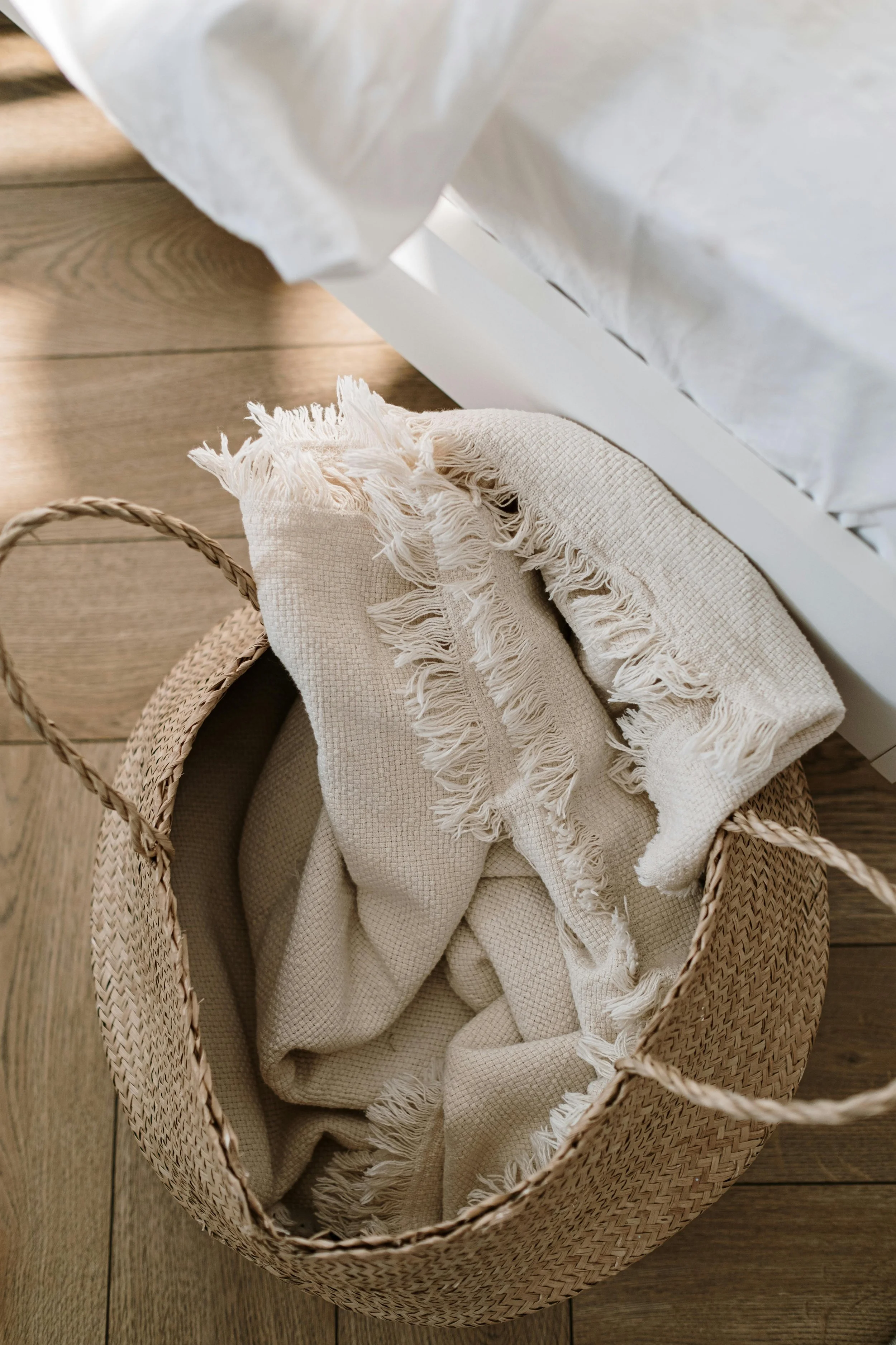 Top-down view of a cream-colored textured fringe throw blanket inside a natural woven seagrass basket on a light oak wood floor, organic modern home decor aesthetic.