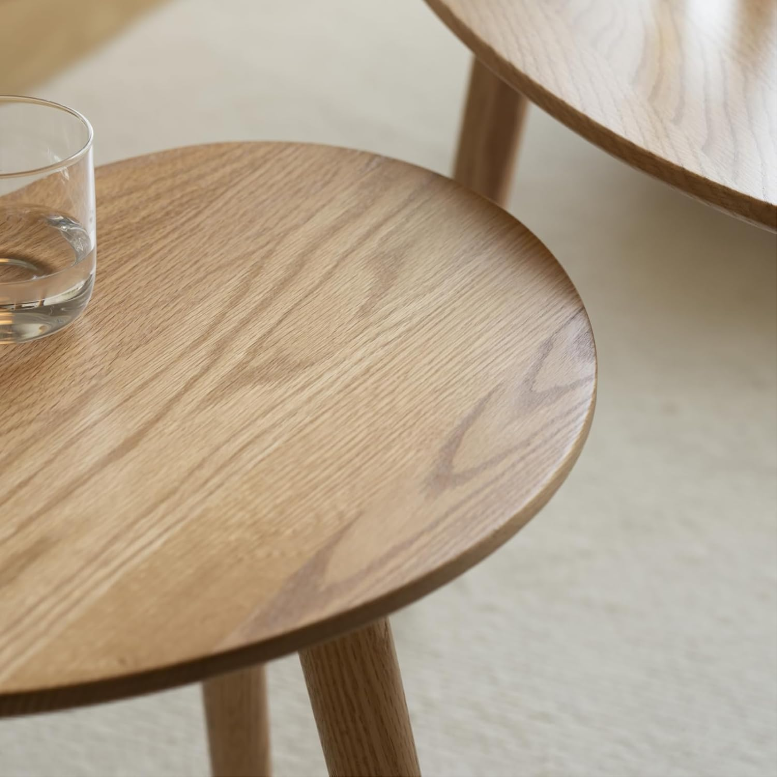 Close-up of a round natural oak nesting coffee table with visible wood grain, featuring a glass of water, styled on a neutral cream rug in a minimalist living room.