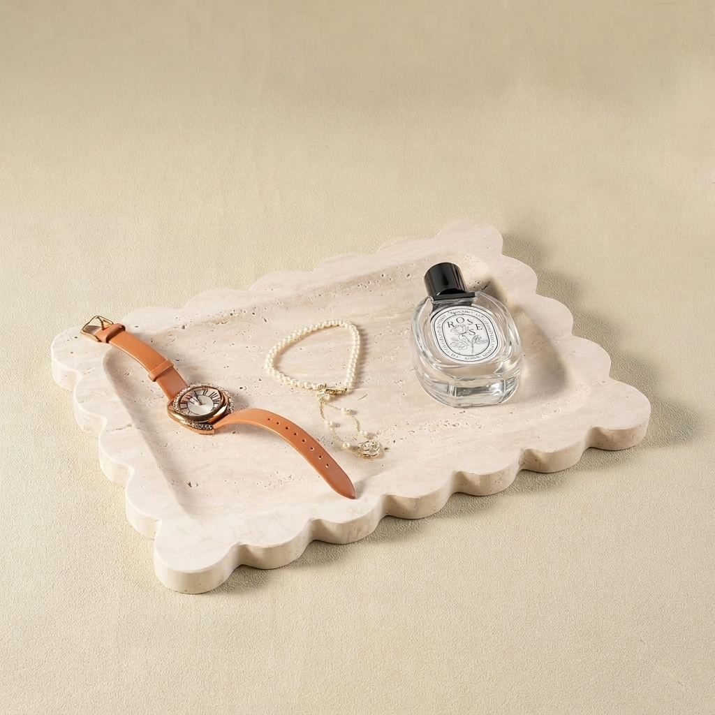 A scalloped-edge tray made of genuine beige travertine stone, styled with a leather watch, a pearl bracelet, and a bottle of Diptyque Rose perfume on a neutral cream background.