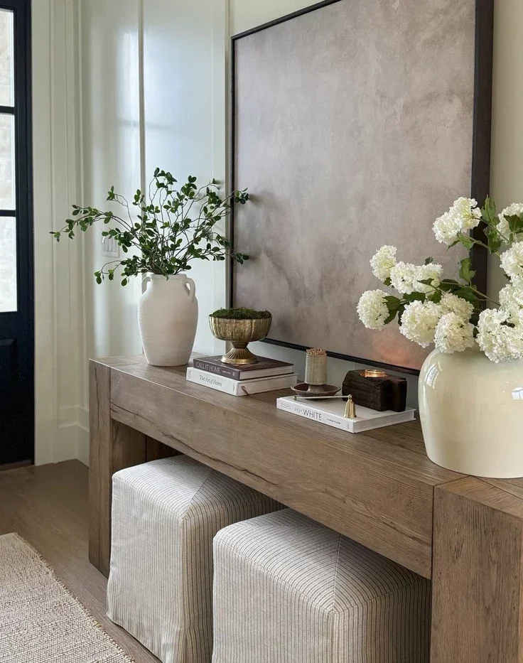 A rustic wooden console table in an entryway styled with a large beige abstract painting, a white vase filled with white hydrangeas, and two neutral striped fabric ottoman cubes tucked underneath.