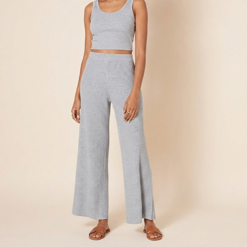 A full-body photograph of a woman with dark curly hair wearing a matching grey ribbed knit sleeveless crop top and high-waisted, wide-leg pants set, wearing brown flat sandals.