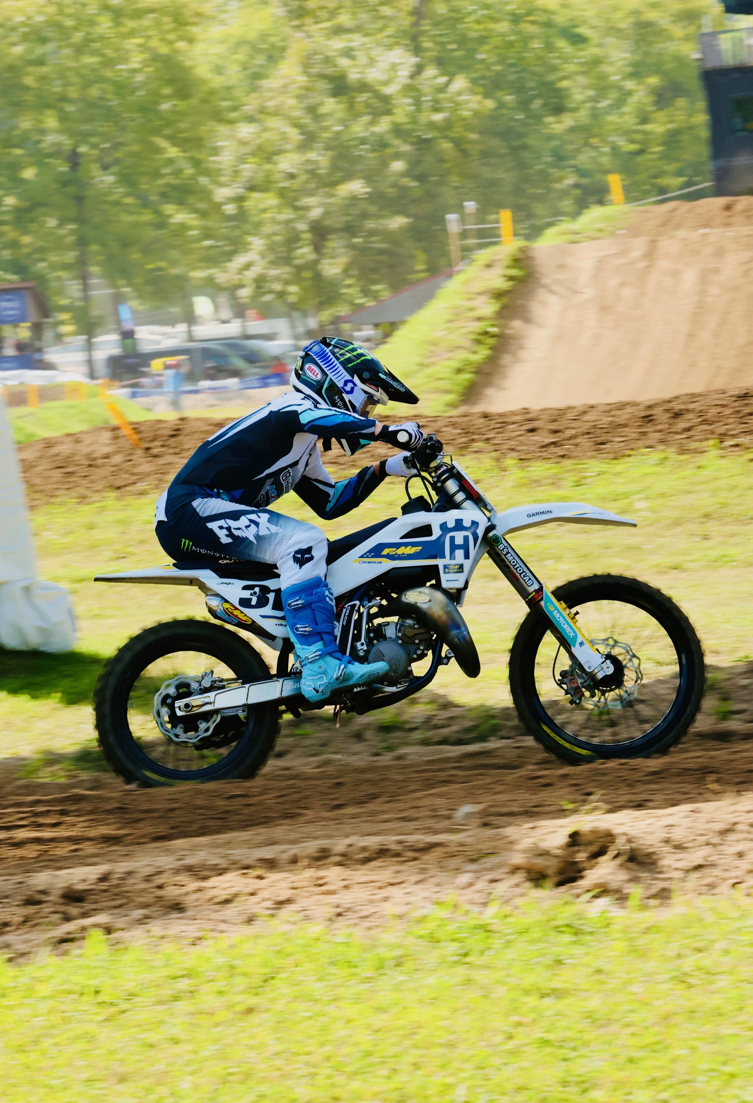Motorcycle rider wearing blue and black gear, helmet, and gloves, racing on a dirt track with trees and vehicles in background.