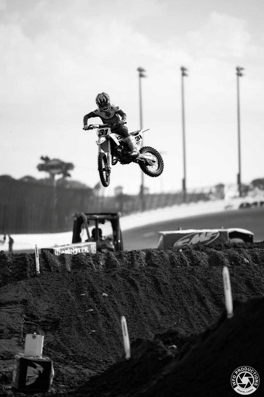 A motocross rider in mid-air during a jump on a dirt track, with flags, construction equipment, and trees in the background, captured in black and white.