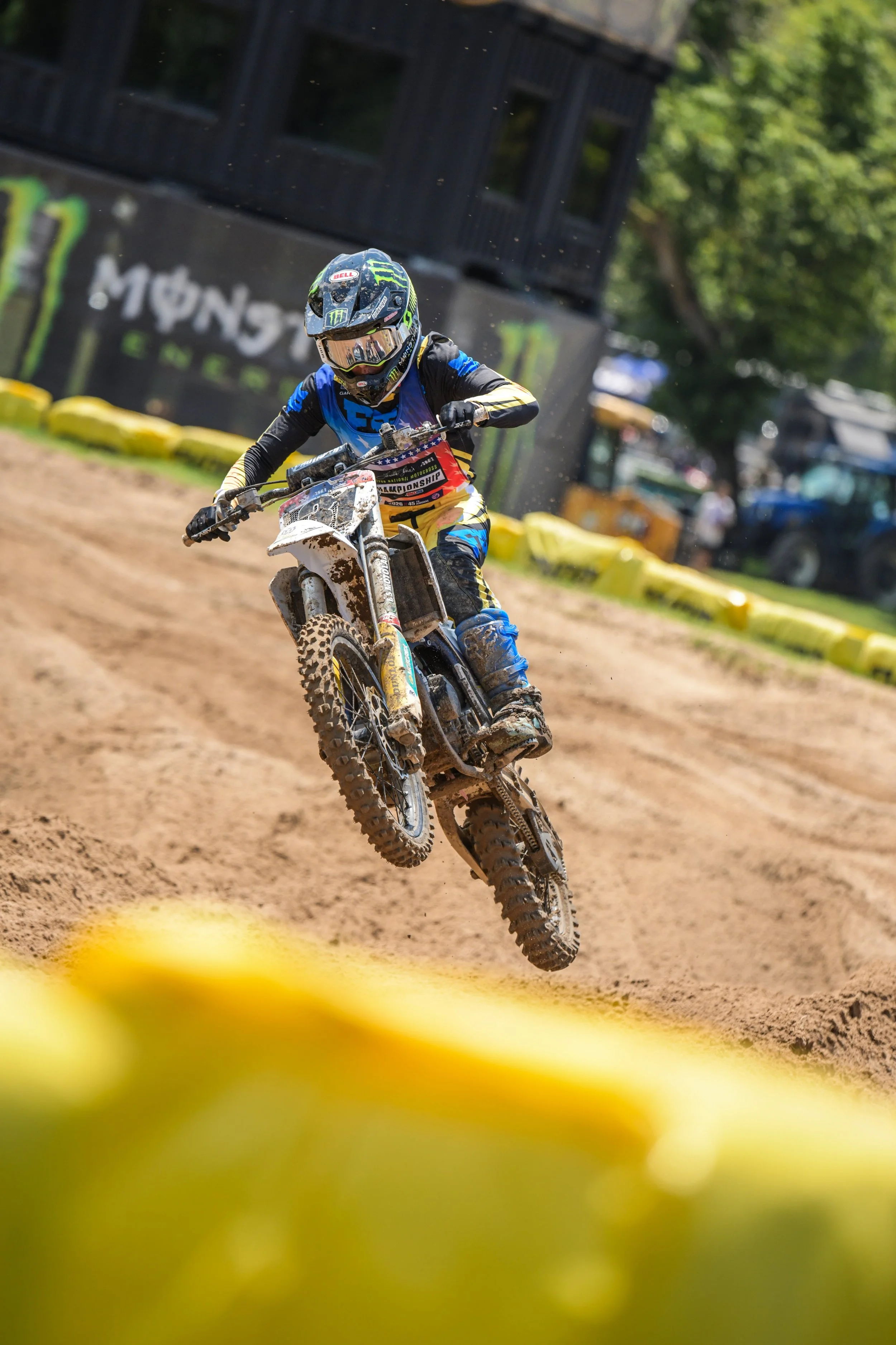 A motocross rider in a blue, black, and yellow suit and helmet riding a dirt bike on a dirt track during a race. The rider is airborne, and the background shows trees, a black structure, and some yellow barriers.