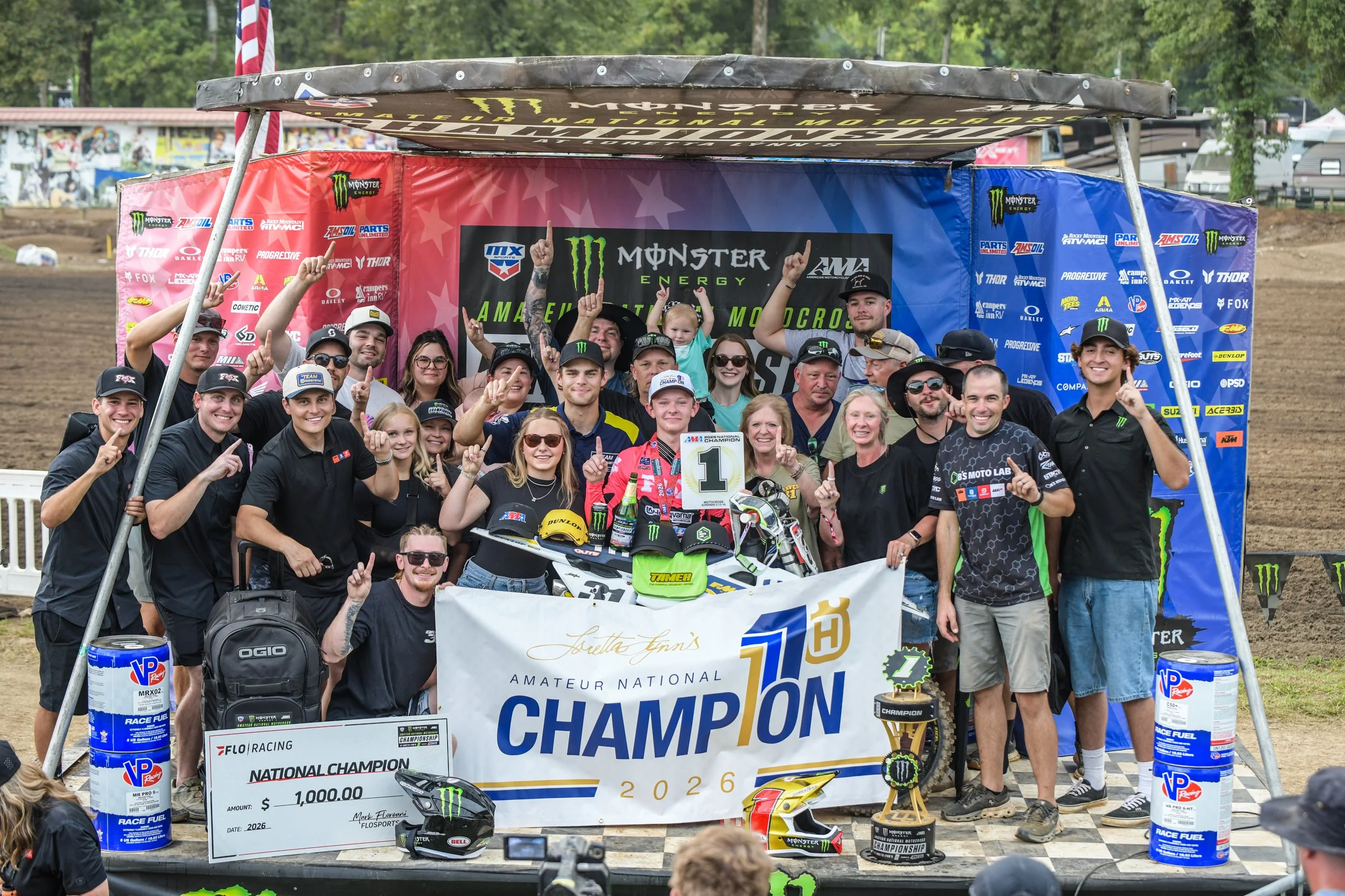 A group of people, including racers and supporters, celebrating at a motocross event, holding a large sign with a prize check for $1,000, surrounded by banners and motocross gear.