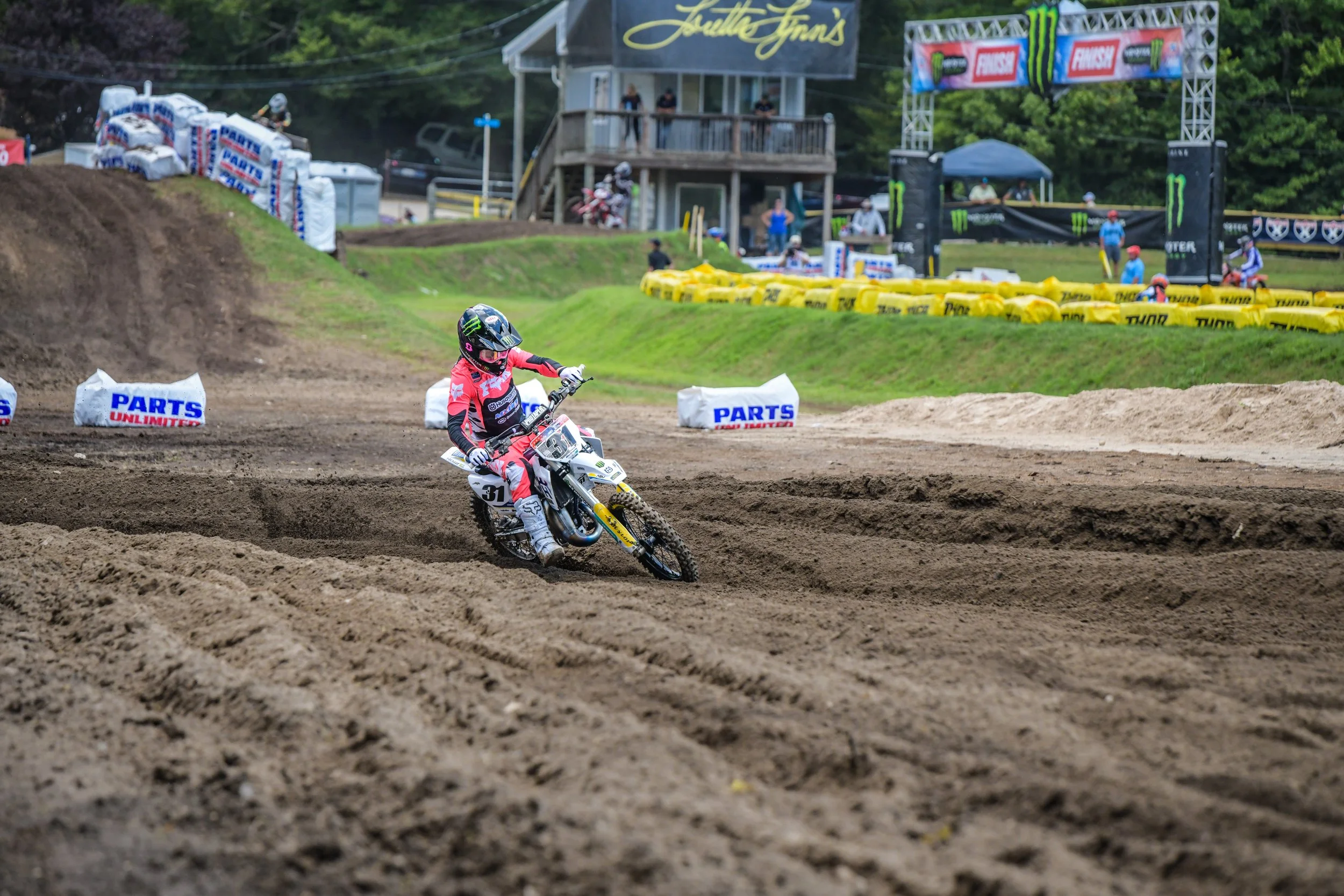 A dirt bike rider wearing a pink racing suit and helmet is navigating a dirt track at a motocross event. The background features spectators, yellow barriers, advertising signs, and a structure with staff and riders.