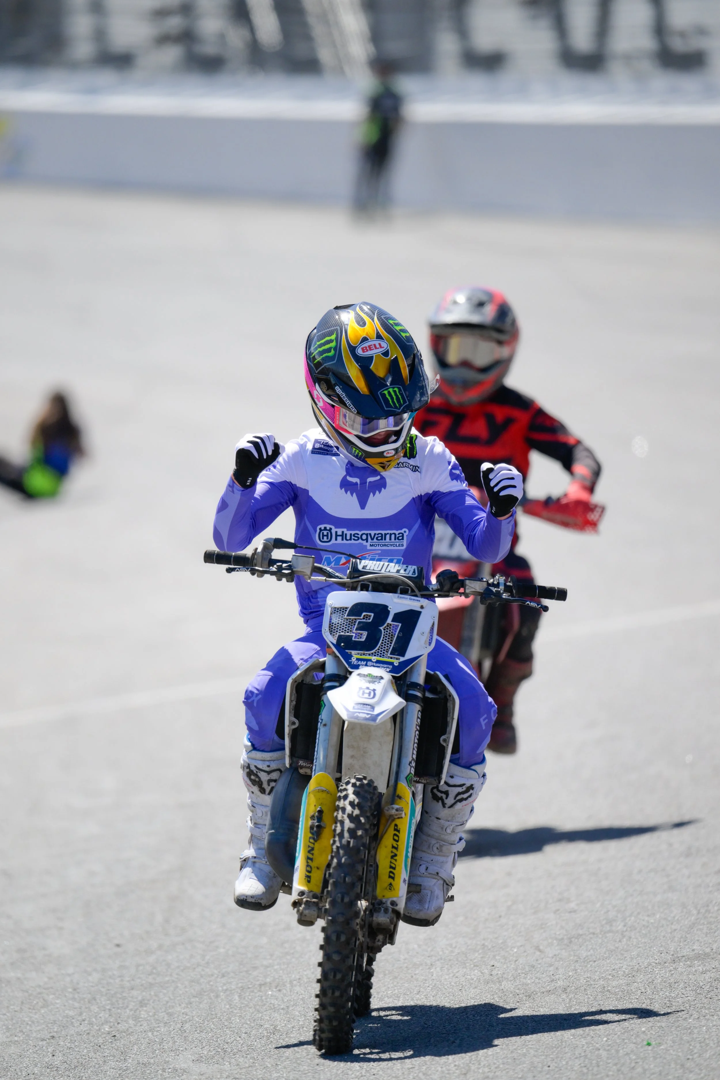 A young motocross rider in blue gear pedaling a dirt bike on a track, with other motorcyclists and spectators blurred in the background.