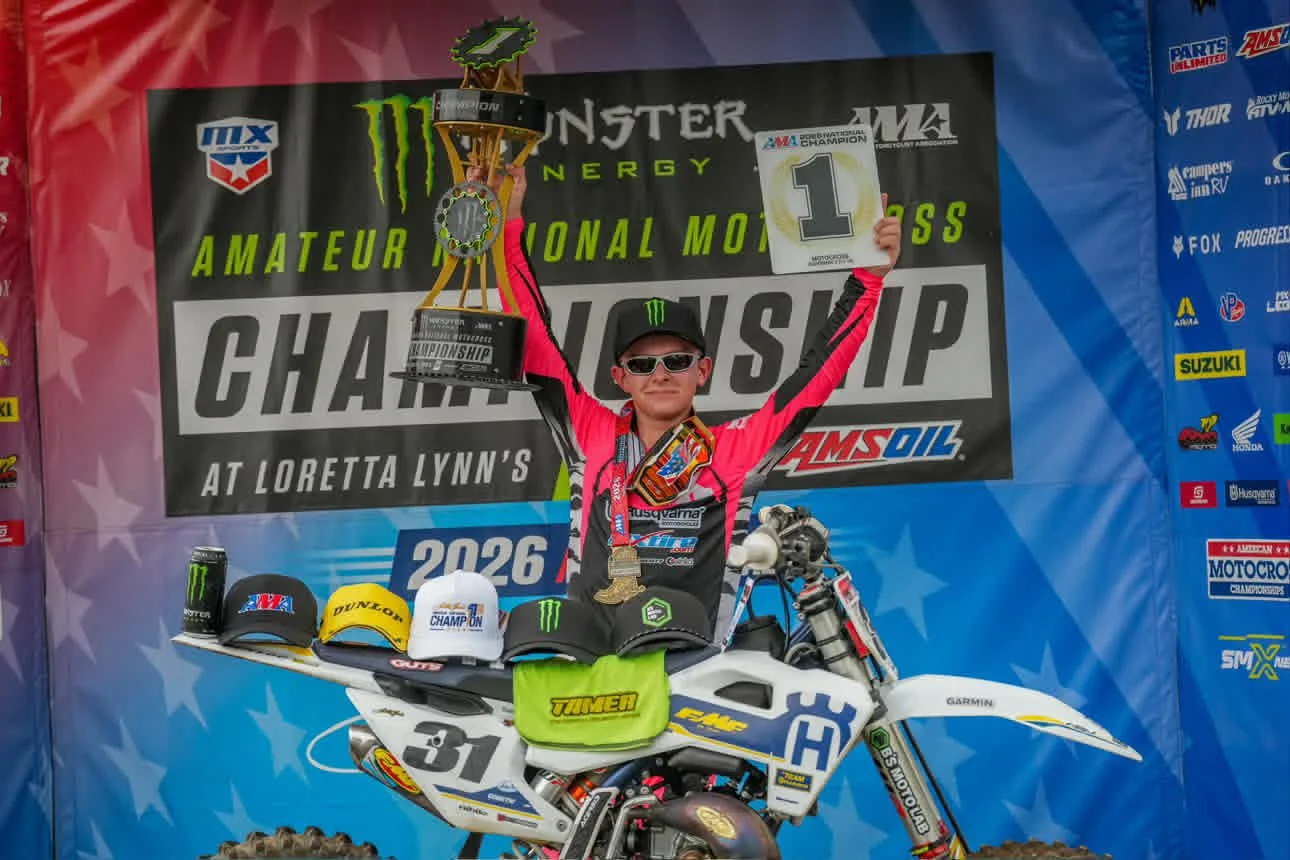 Young motocross rider celebrates on podium holding a trophy and a first-place sign, standing behind his dirt bike with multiple racing hats displayed on the bike's front fender, in front of a championship backdrop.