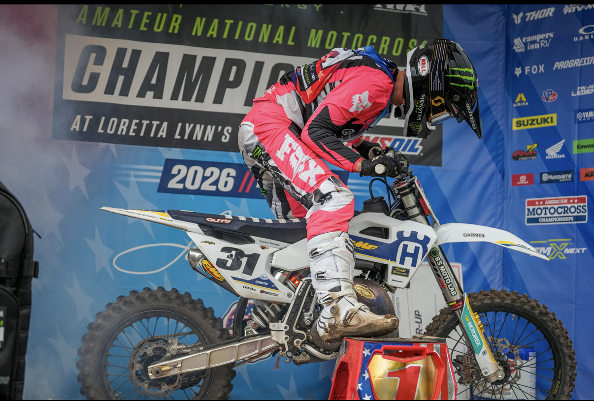 A motocross rider in a pink and black racing suit and helmet, standing on a white Husqvarna dirt bike, preparing for a race at the 2026 Amateur National Motocross Championship, with a backdrop displaying race details and sponsors.