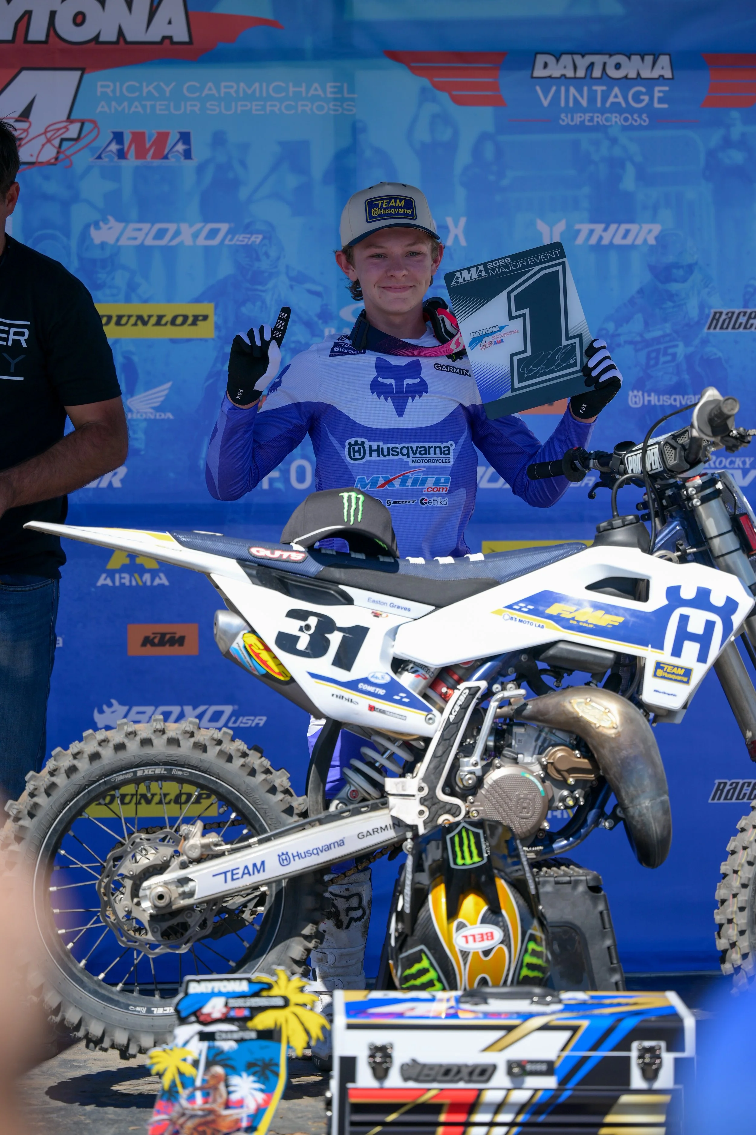 A young man in motocross gear standing next to a dirt bike, holding a winner's certificate at a racing event. He is smiling and making a peace sign. The background features racing banners and logos.