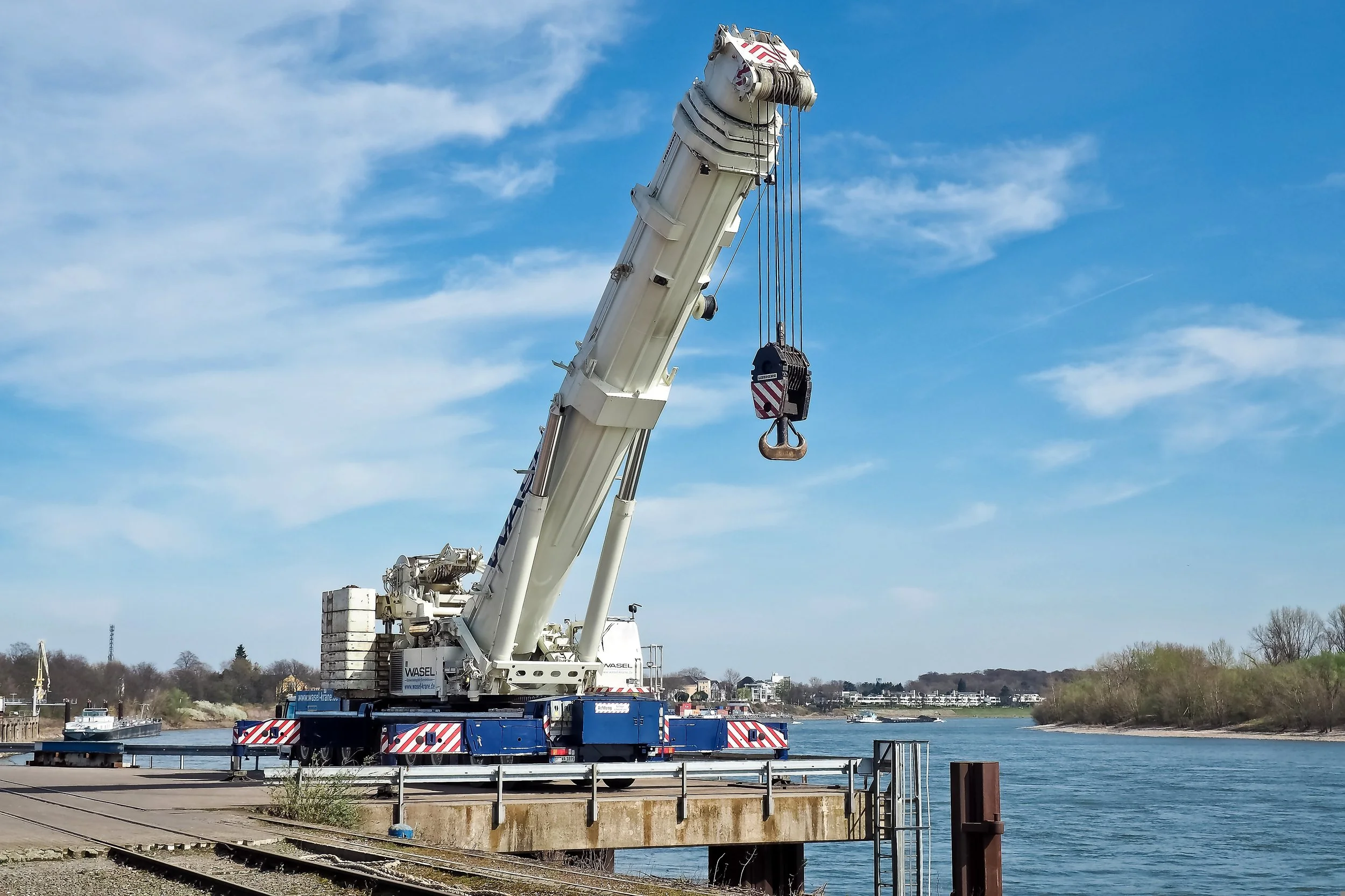 A large telescoping crane with a white body and red warning stripes on the side, positioned near a river, with its arm extended upward. The crane's hook is hanging down. The scene includes a calm water body, a clear blue sky with some clouds, trees, and buildings in the background.