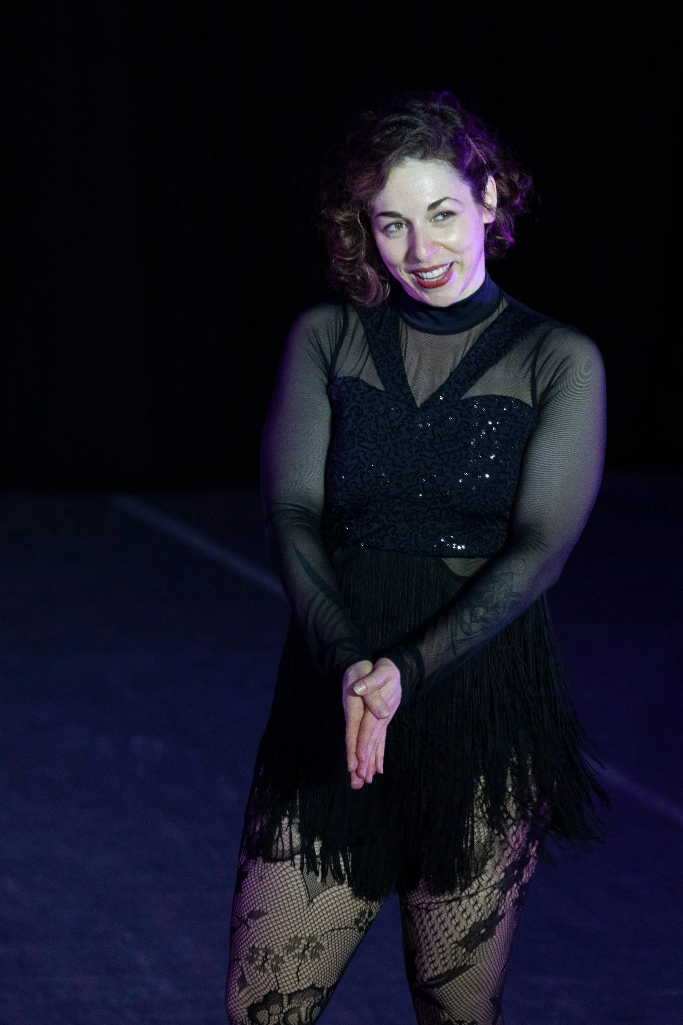 A woman in a black mesh, sequin, and fringe outfit speaks to the audience.