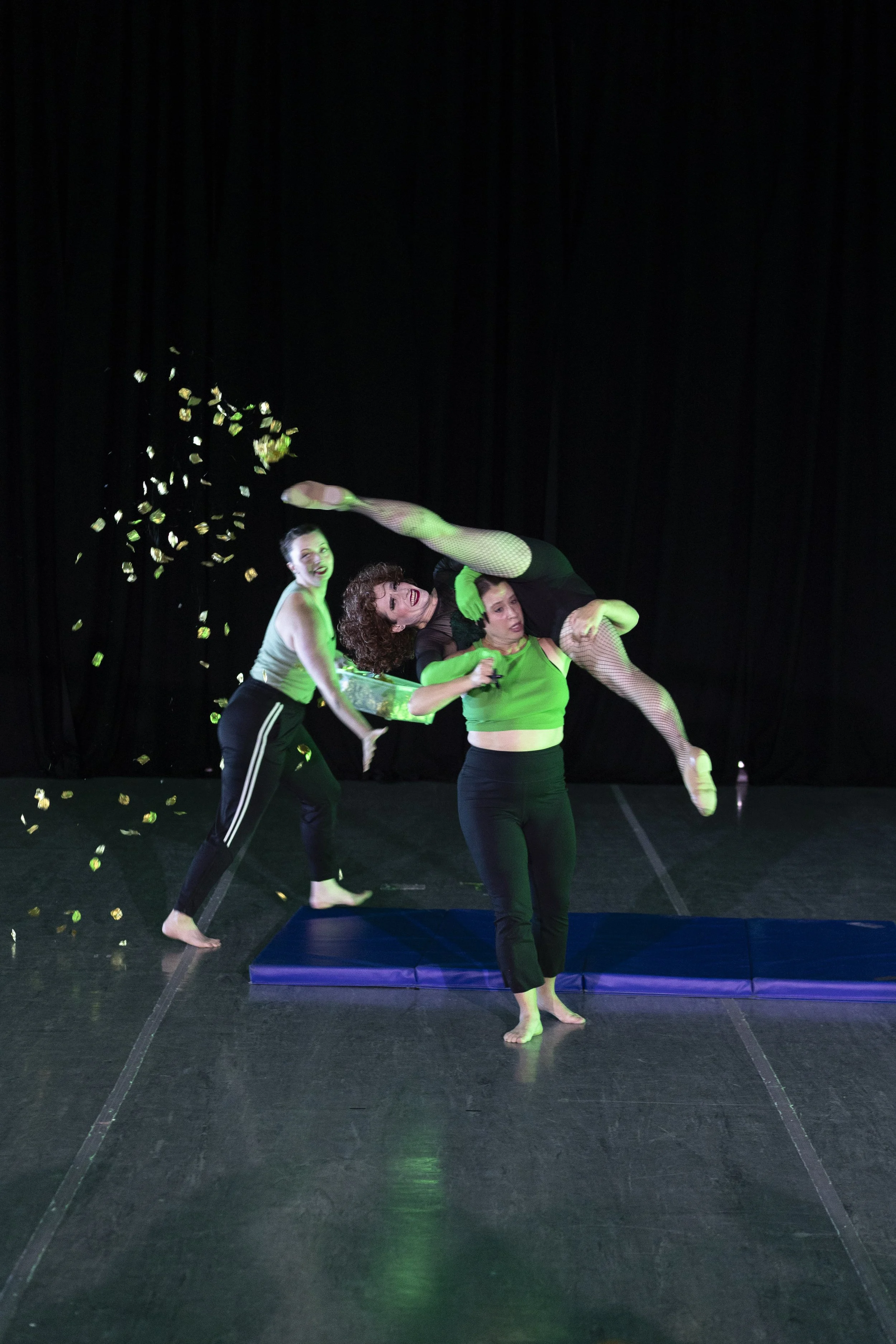 Dancers in green and black perform. One dancer lifts another, face looking strained. Another walks by and throws gold rose petals into the air.