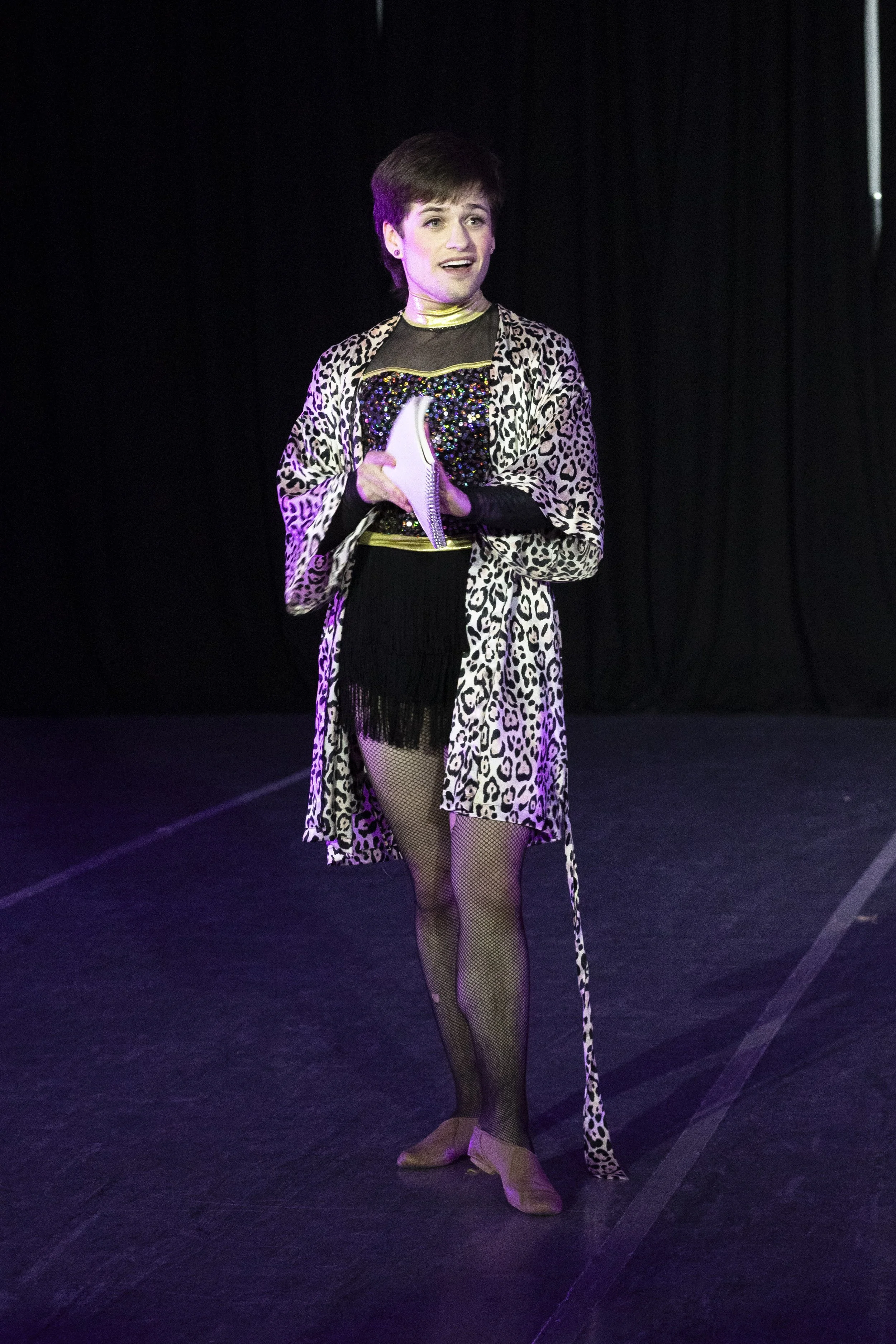 A person in a gold and black mesh, fringe, and sequin outfit wears a leopard print silk robe and holds papers, addressing the audience.