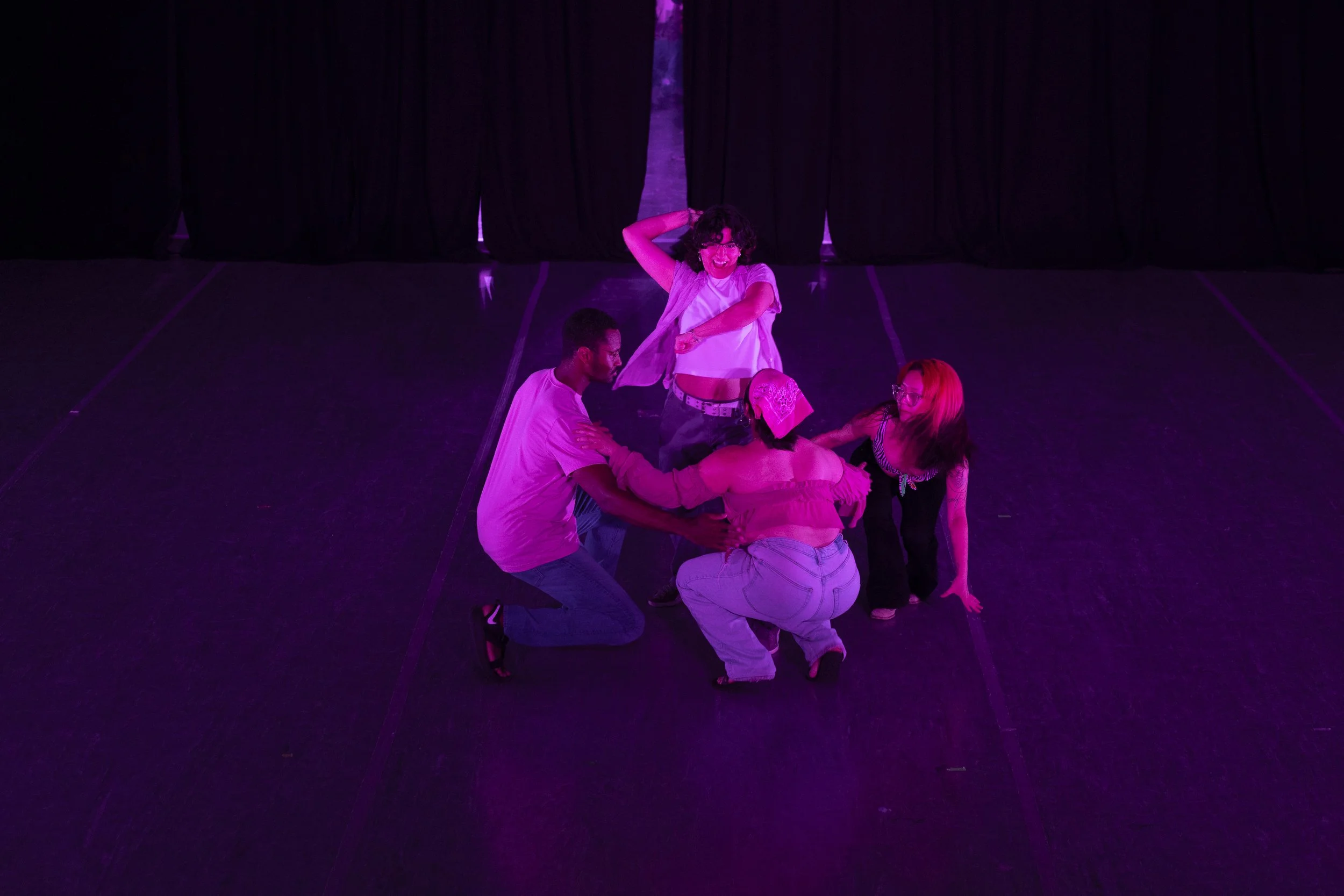 A group of dancers wearing pink and blue perform onstage. Four dancers crouch in a circle around a dancer who stands up and does a lasso motion.
