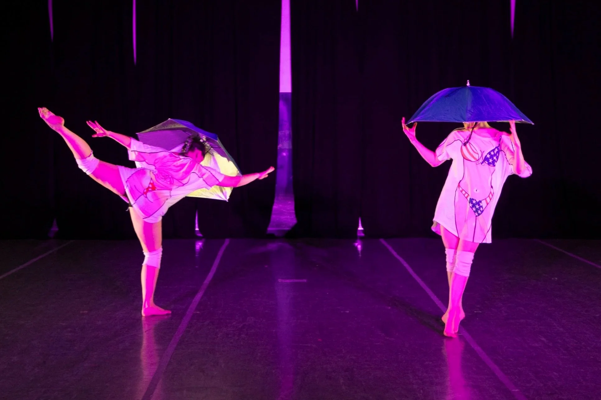 Two dancers wear t-shirts with bodies wearing American flag bikinis on them. They wear umbrella hats and knees pads. One is doing an arabesque.
