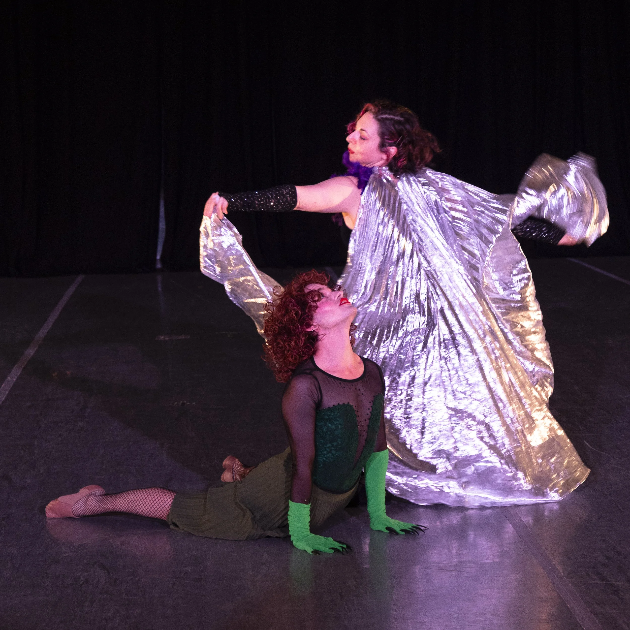 Miss Vikki and Miss Victoria hit poses onstage. Miss Victoria's silver cape is outstretched as she runs behind Ms. Vikki, who is doing an upward dog pose.