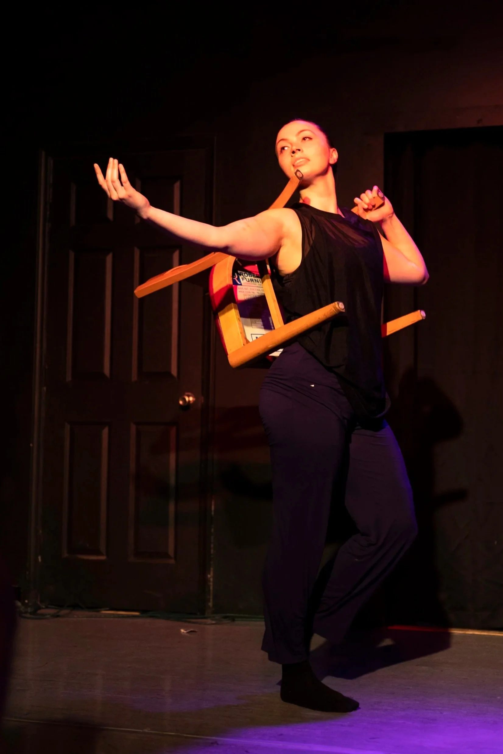 A dancer dressed in all black carries a chair on her back and gestures dramatically toward the audience.