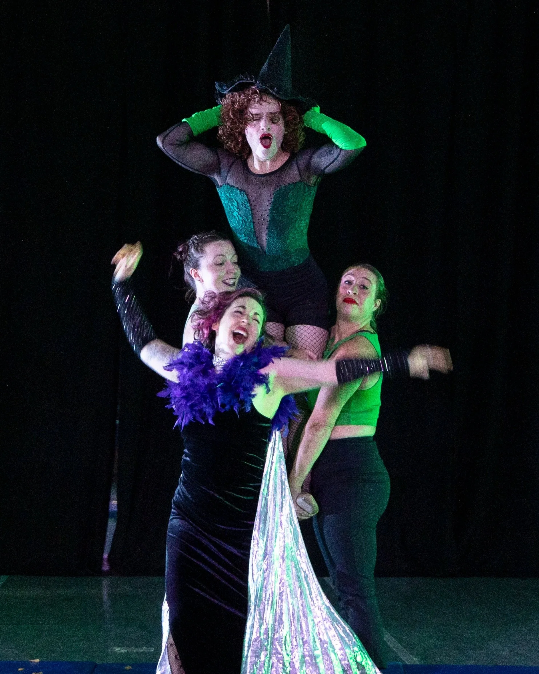 Two dancers lift a dancer who holds a witch hat, singing. Another dancer in a witch costume gestures dramatically in front of them.