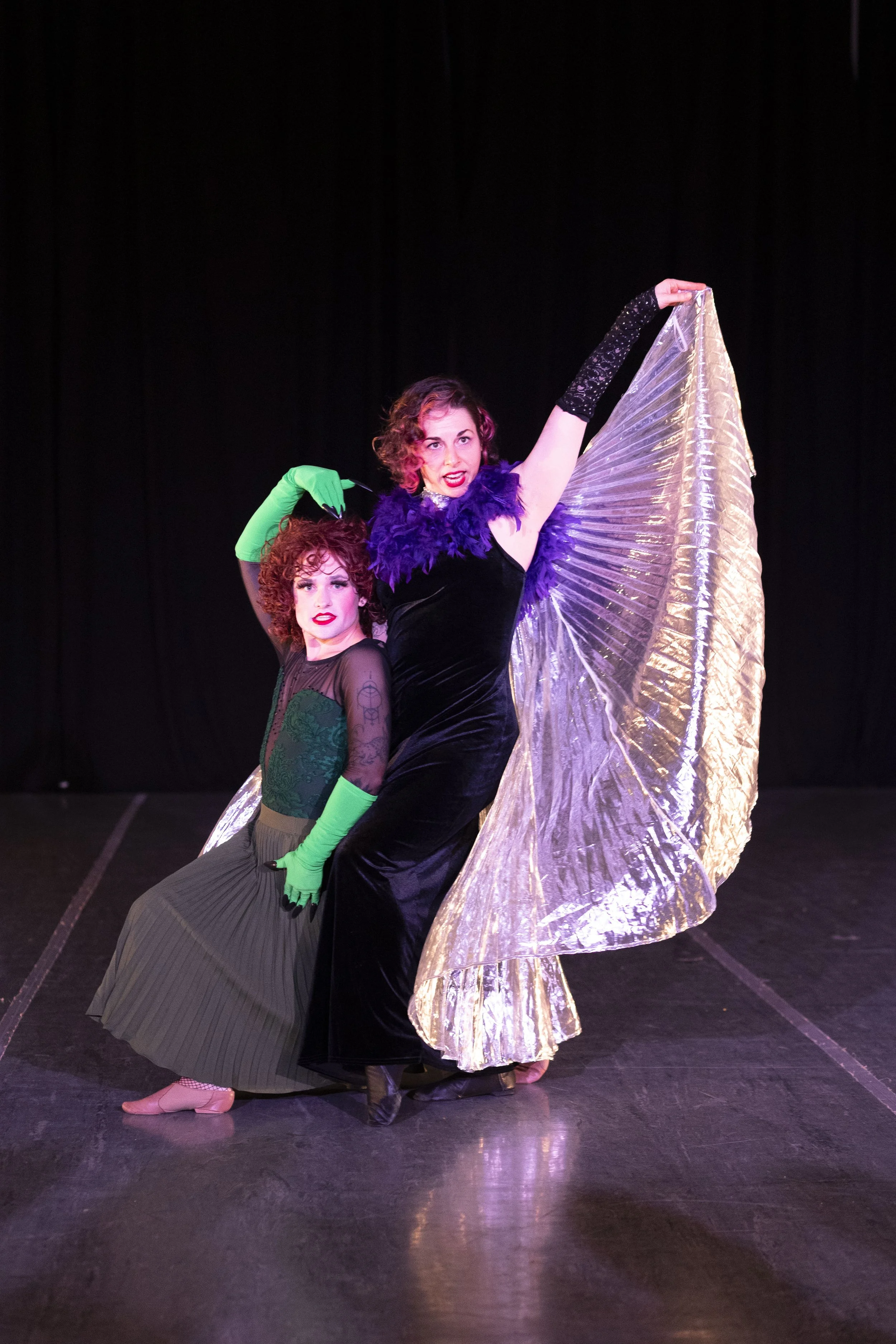 Two women with curly red hair and stage makeup pose, making serious but funny faces. They wear witch-inspired costumes to honor the movie Wicked.
