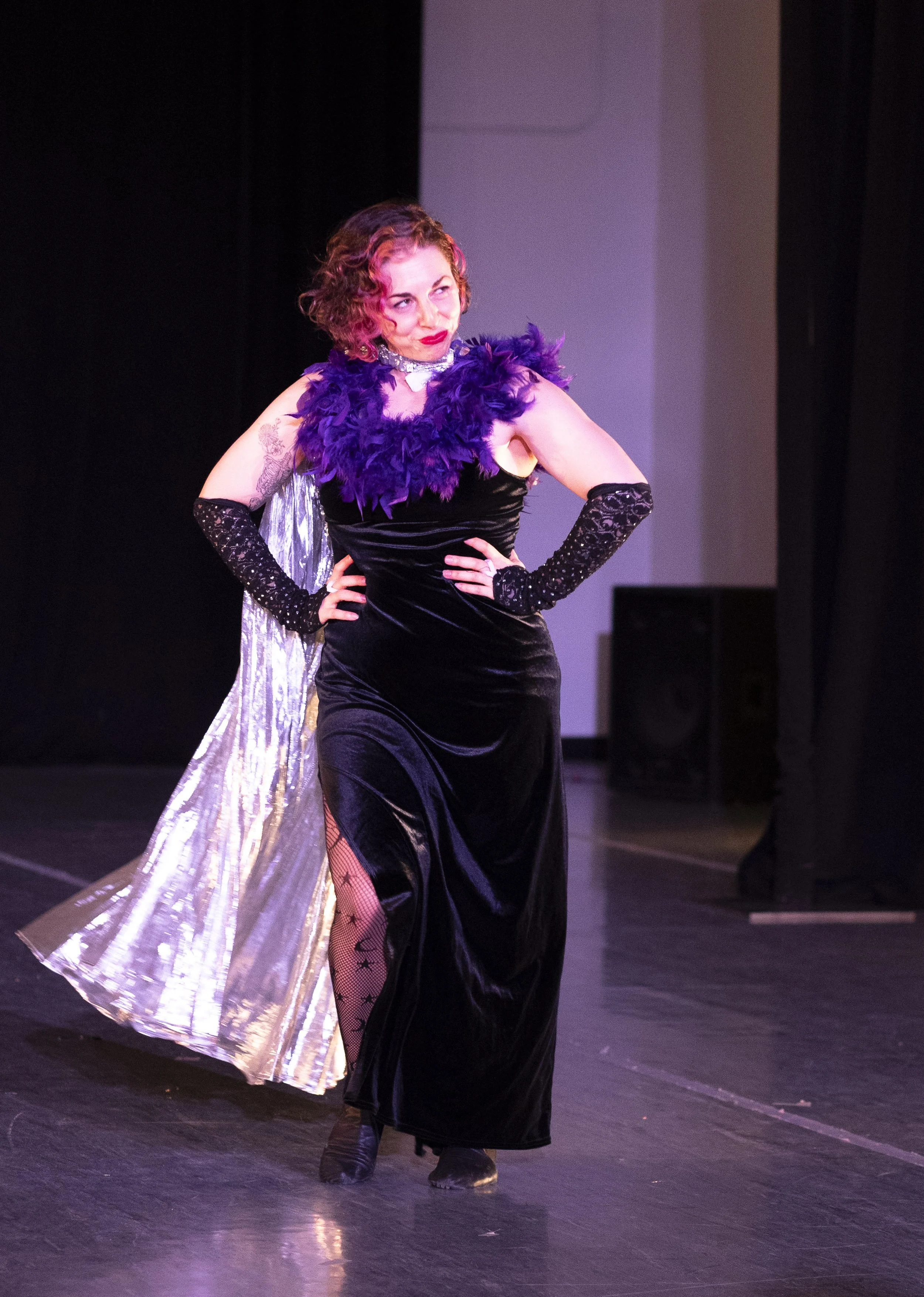 A woman with curly brown & pink hair poses smugly in a long, black velvet dress with a purple feather boa trim. She wears a shiny silver cape.