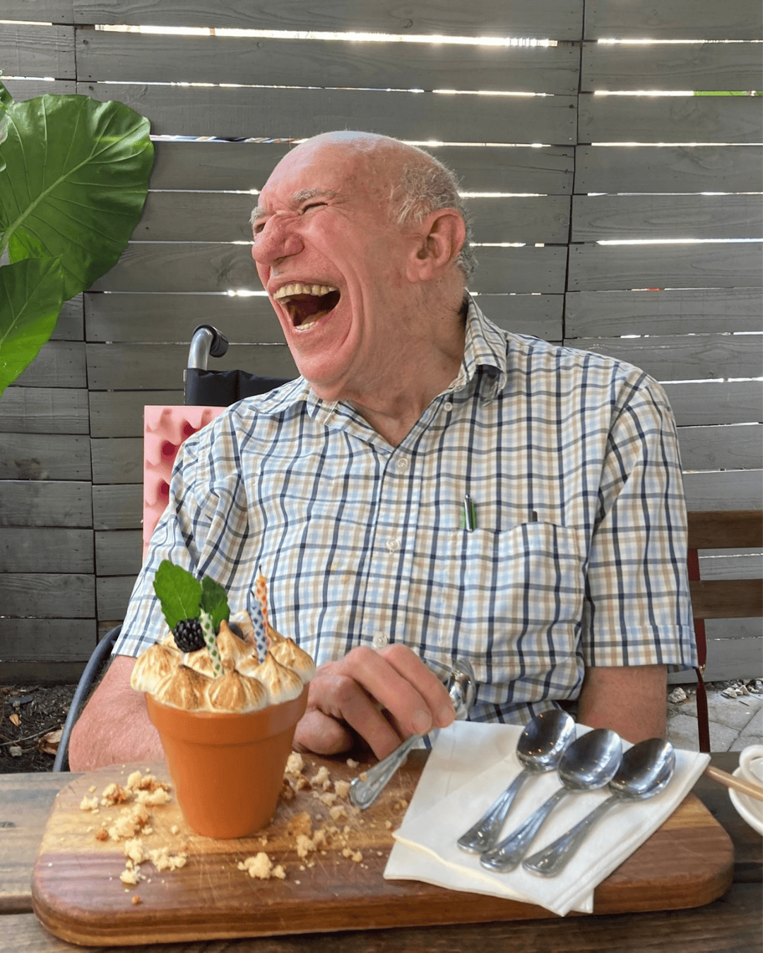 Lolo in a checkered shirt laughing joyfully at Doce Provisiones restaurant in Miami, Florida. On the table is a dessert in a small flowerpot decorated with whipped cream, blackberries, a leaf, and a decorative candle.