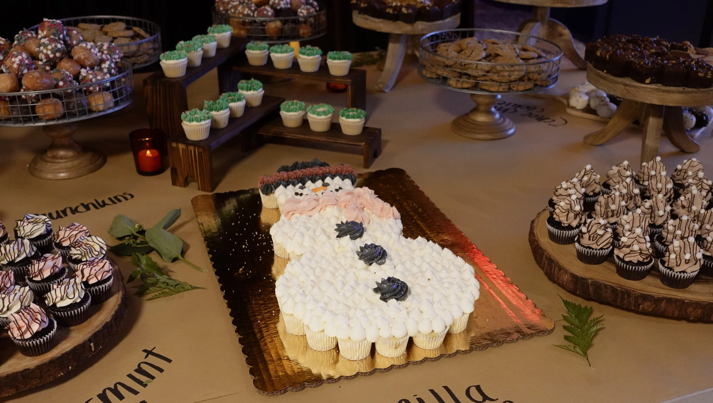 Display of various desserts including cupcakes decorated with green, pink, and black frosting, a large snowman-shaped cake with black and white frosting and black decorative accents, and chocolate chip cookies, all arranged on wooden and metal stands