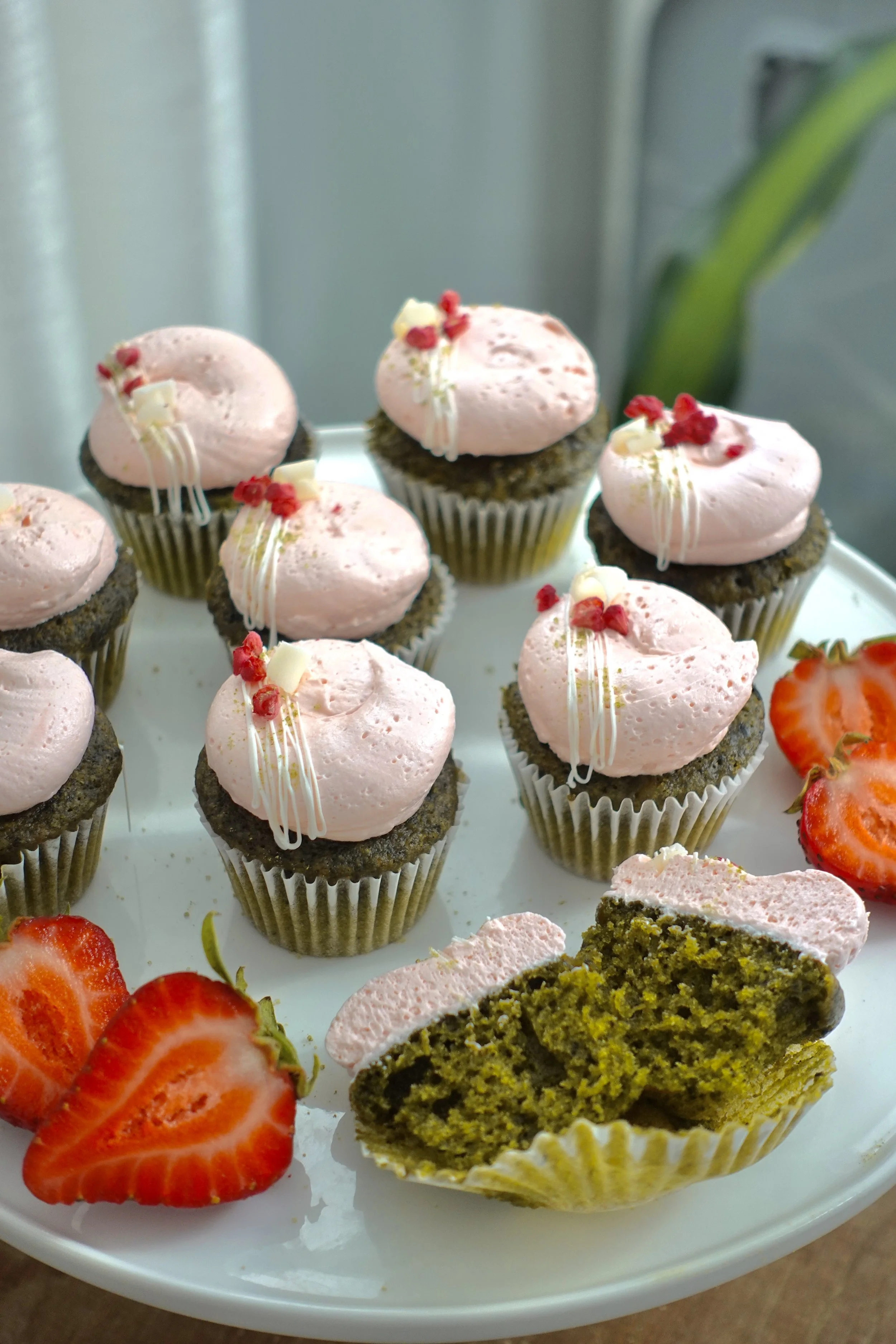 Plate of cupcakes topped with pink frosting, white chocolate decorations, and small red and white edible accents. Sliced strawberries are arranged on the side of the plate.