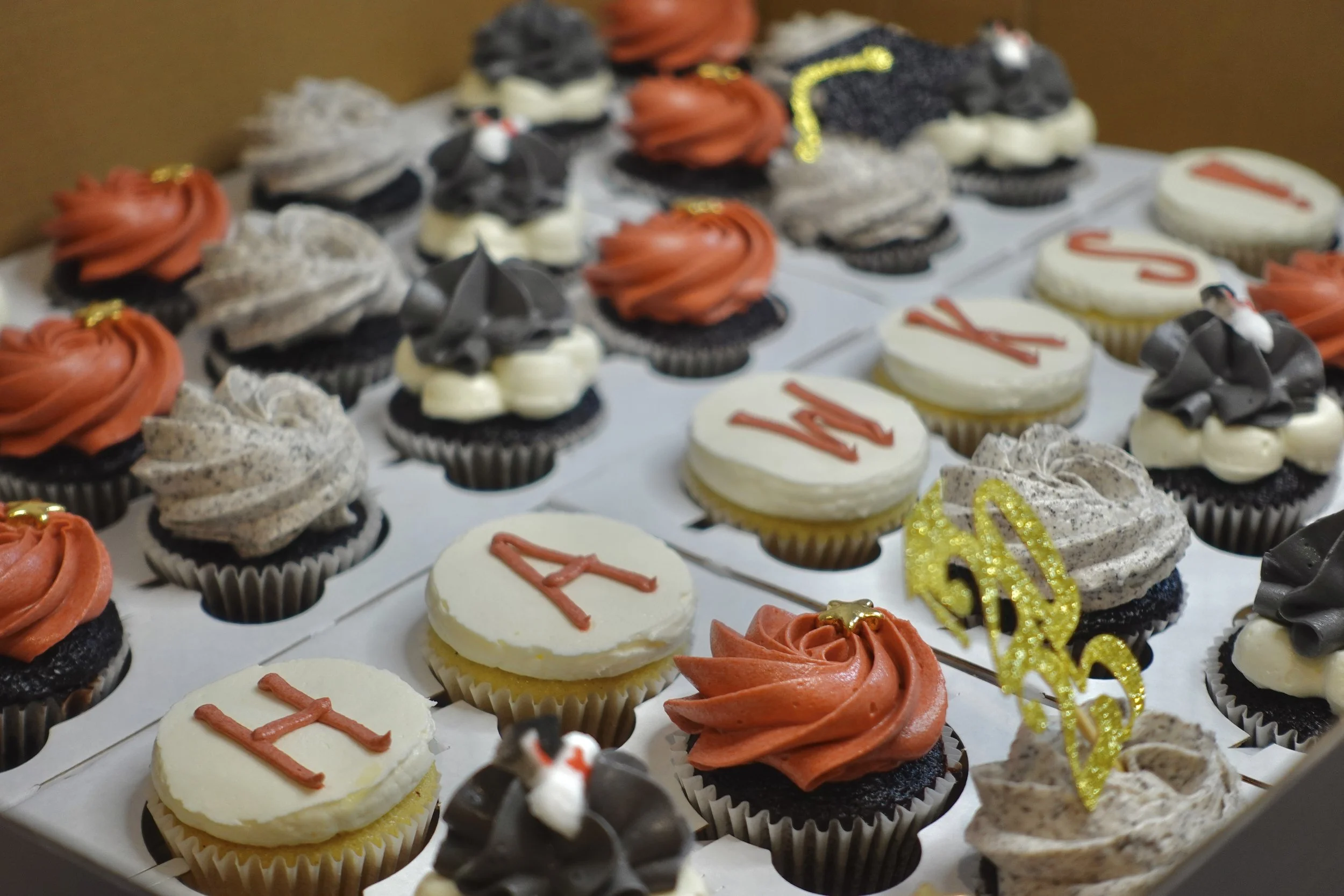 Assorted cupcakes with various frosting designs and decorations, including red, white, black, and gray frosting, some with gold accents, arranged on a baking tray.