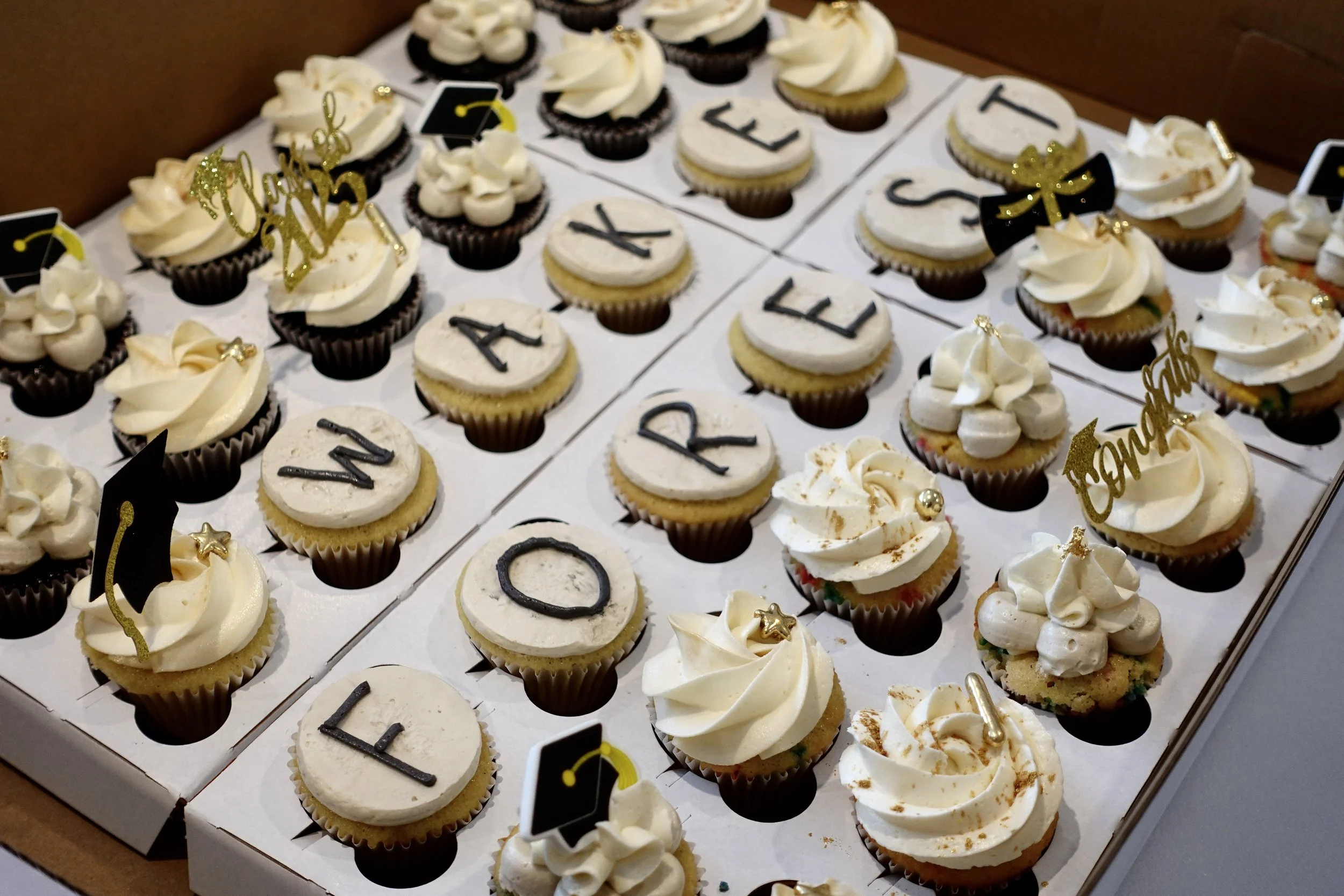 A box filled with assorted cupcakes decorated with white and cream frosting, gold and black decorations, and some topped with edible gold accents. Some cupcakes have letters spelling 'CONGRATULATIONS' and others have small black graduation hat topper