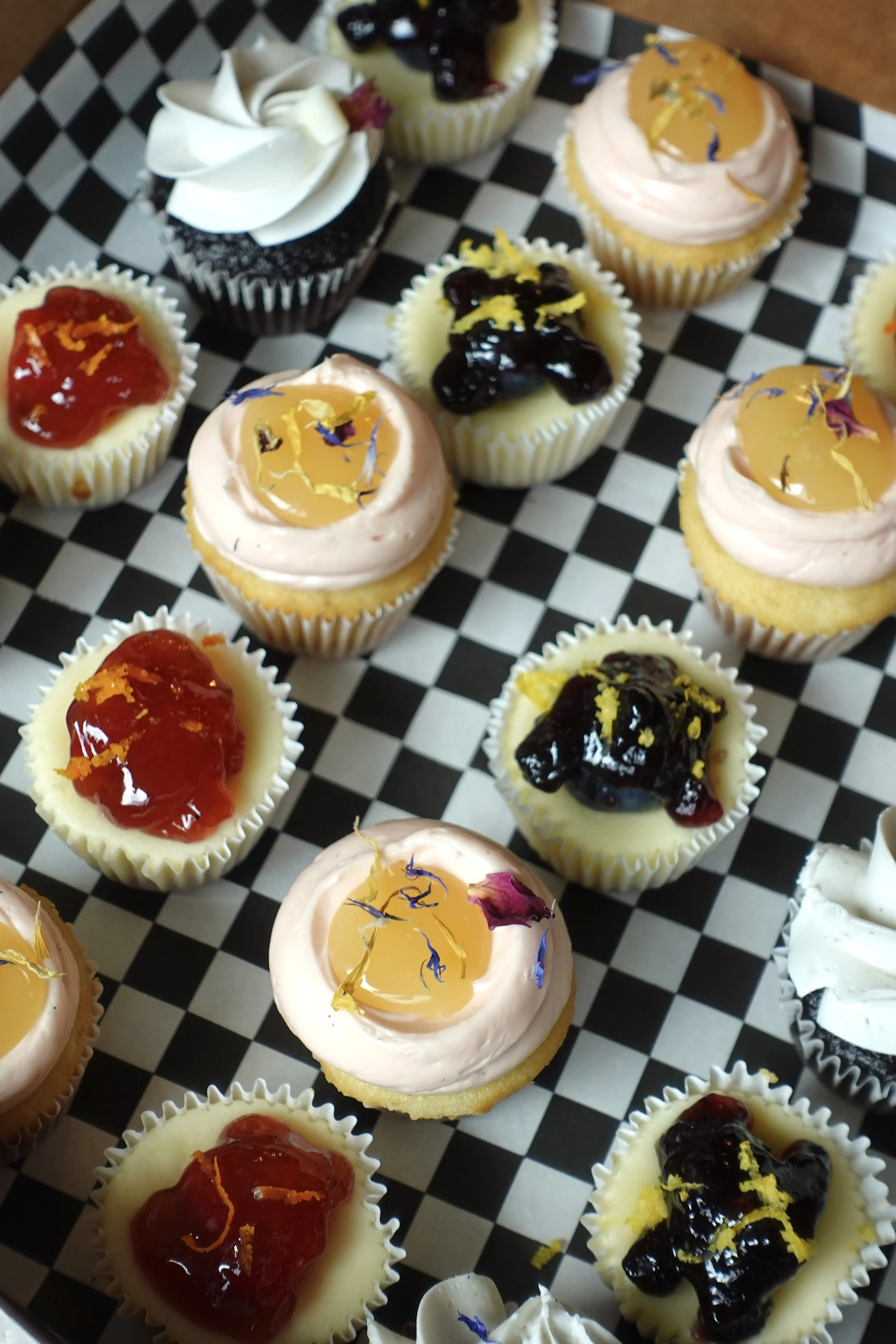 Assorted cupcakes with various toppings including fruit preserves, whipped cream, and edible flower petals, arranged on a black and white checkered surface.