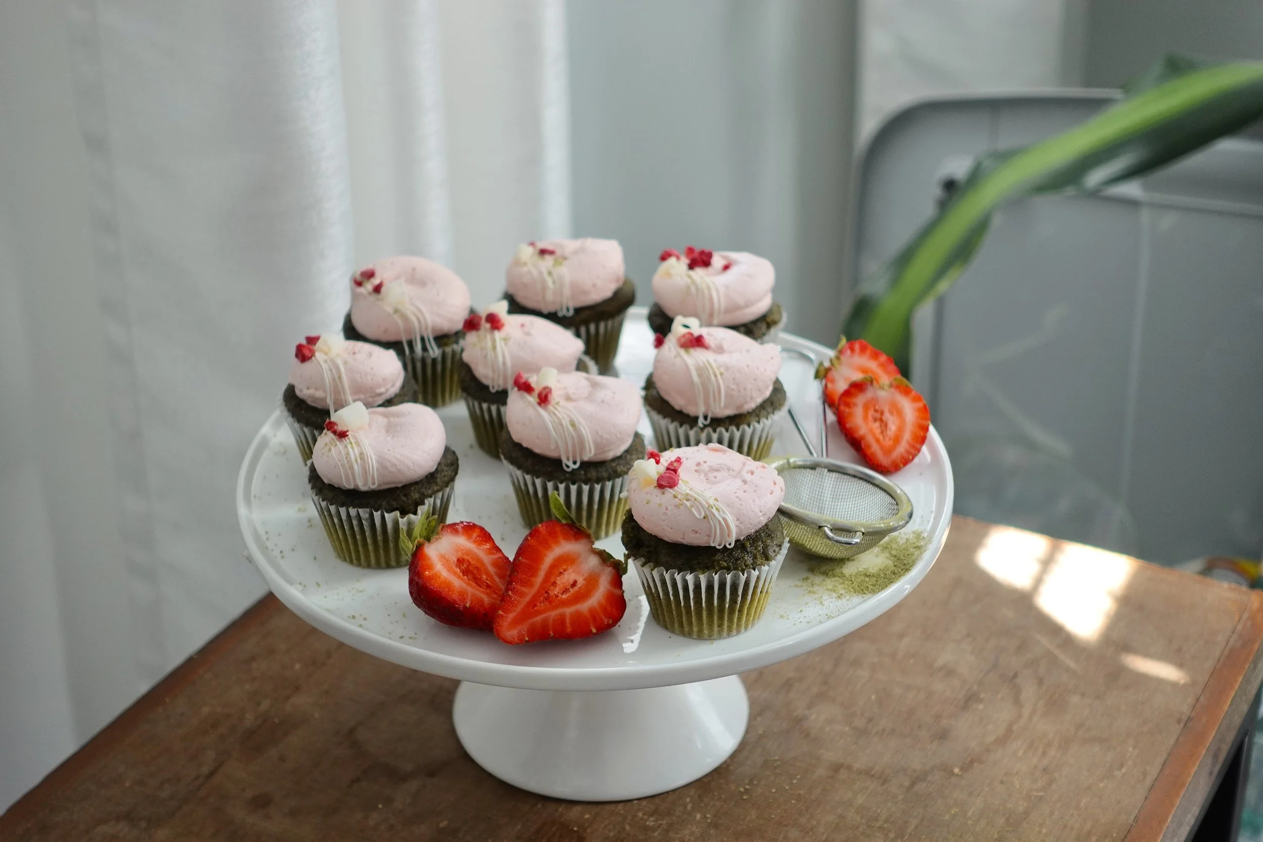 A white cake stand holding chocolate cupcakes with pink frosting, white chocolate decorations, and small red and white edible flowers. Sliced strawberries are on the side of the stand.