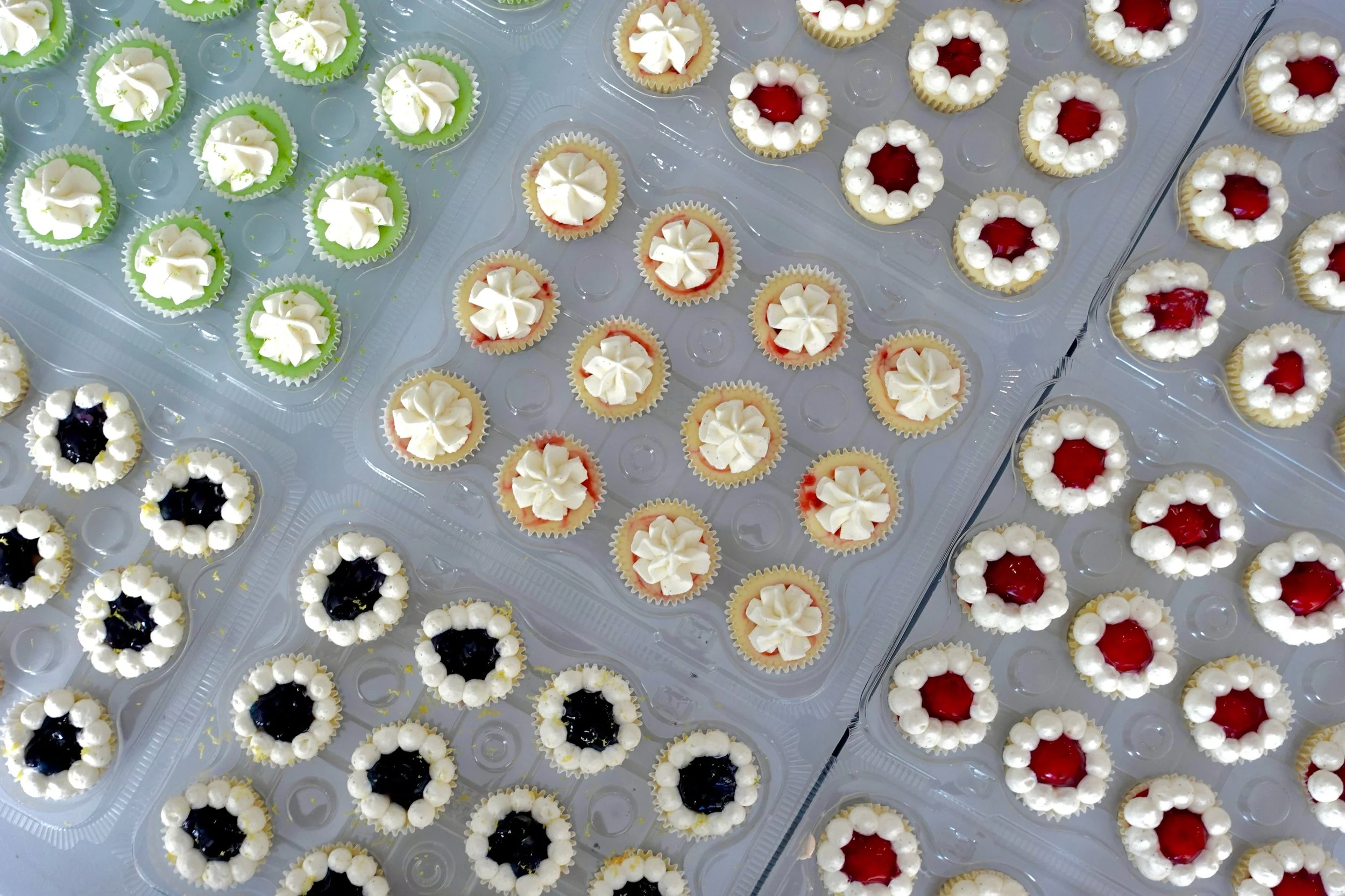 Assorted small cupcakes with white icing, some with red or dark purple fruit topping, arranged in clear plastic trays.