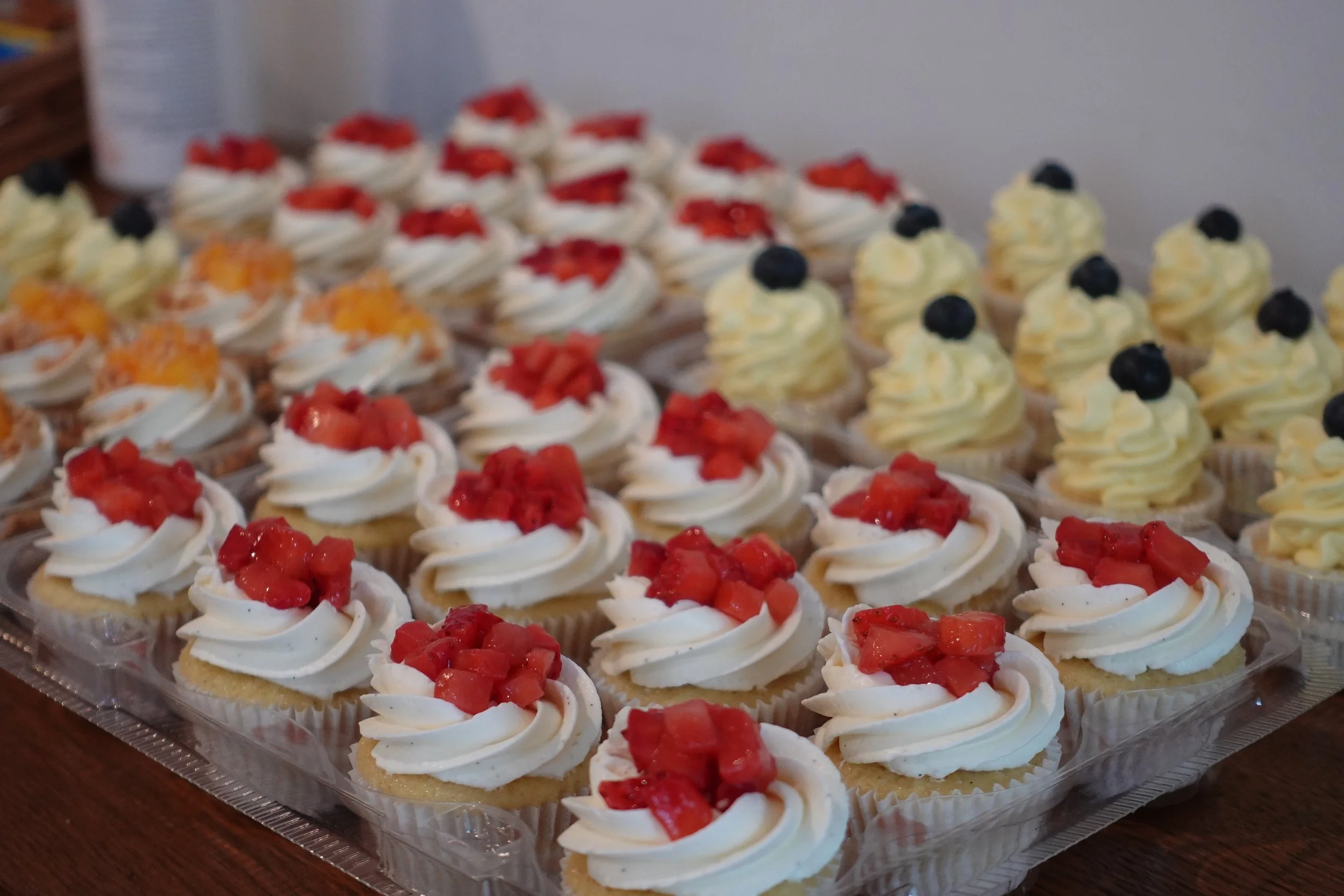 Tray of assorted cupcakes topped with whipped cream and decorated with strawberries, blueberries, and fruit chunks.