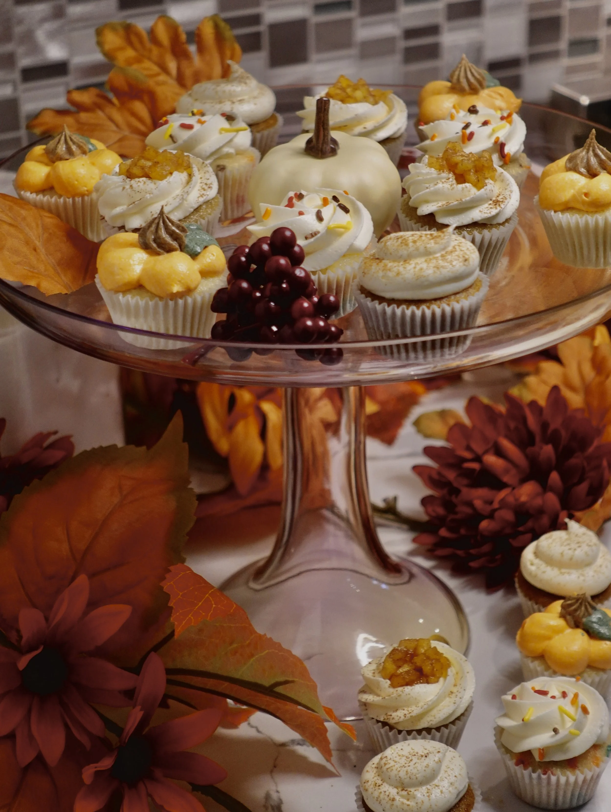 An assortment of cupcakes decorated with various toppings on a glass cake stand, surrounded by autumn leaves and flowers.