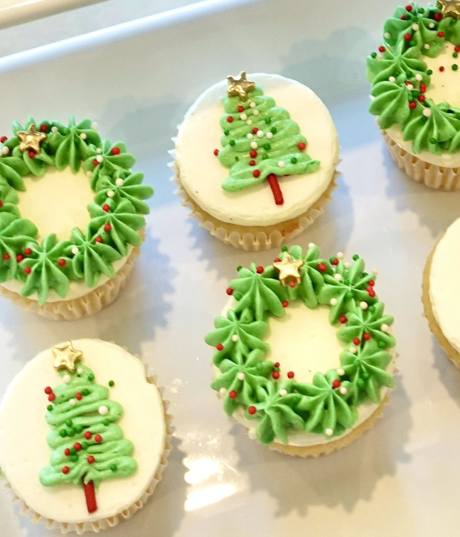 Cupcakes decorated with green frosting piped to resemble Christmas trees, topped with small red, white, and green sprinkles and gold star toppers, on a silver tray.
