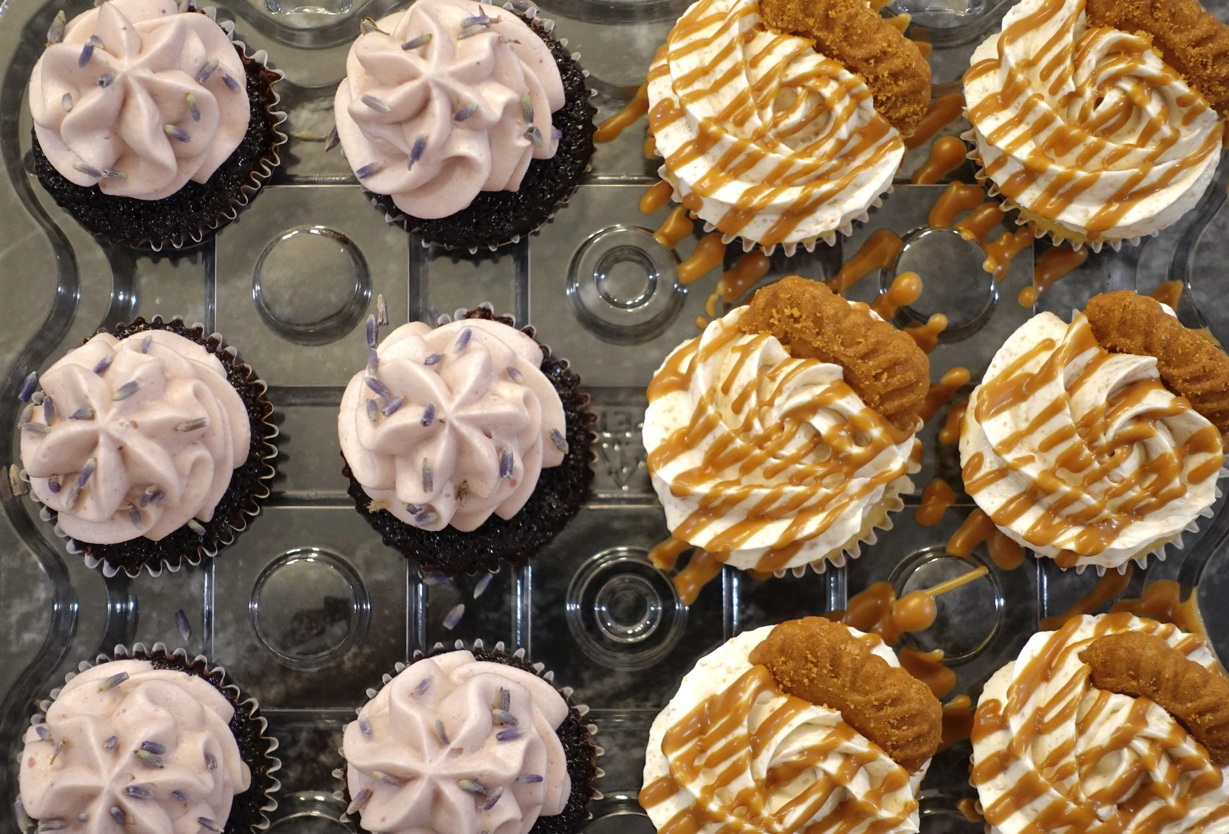 Assorted cupcakes with pink frosting and lavender sprinkles on the left, topped with black cupcake liners; on the right, cupcakes with white frosting, caramel drizzle, and fried chicken strips on top, displayed on a clear plastic tray.