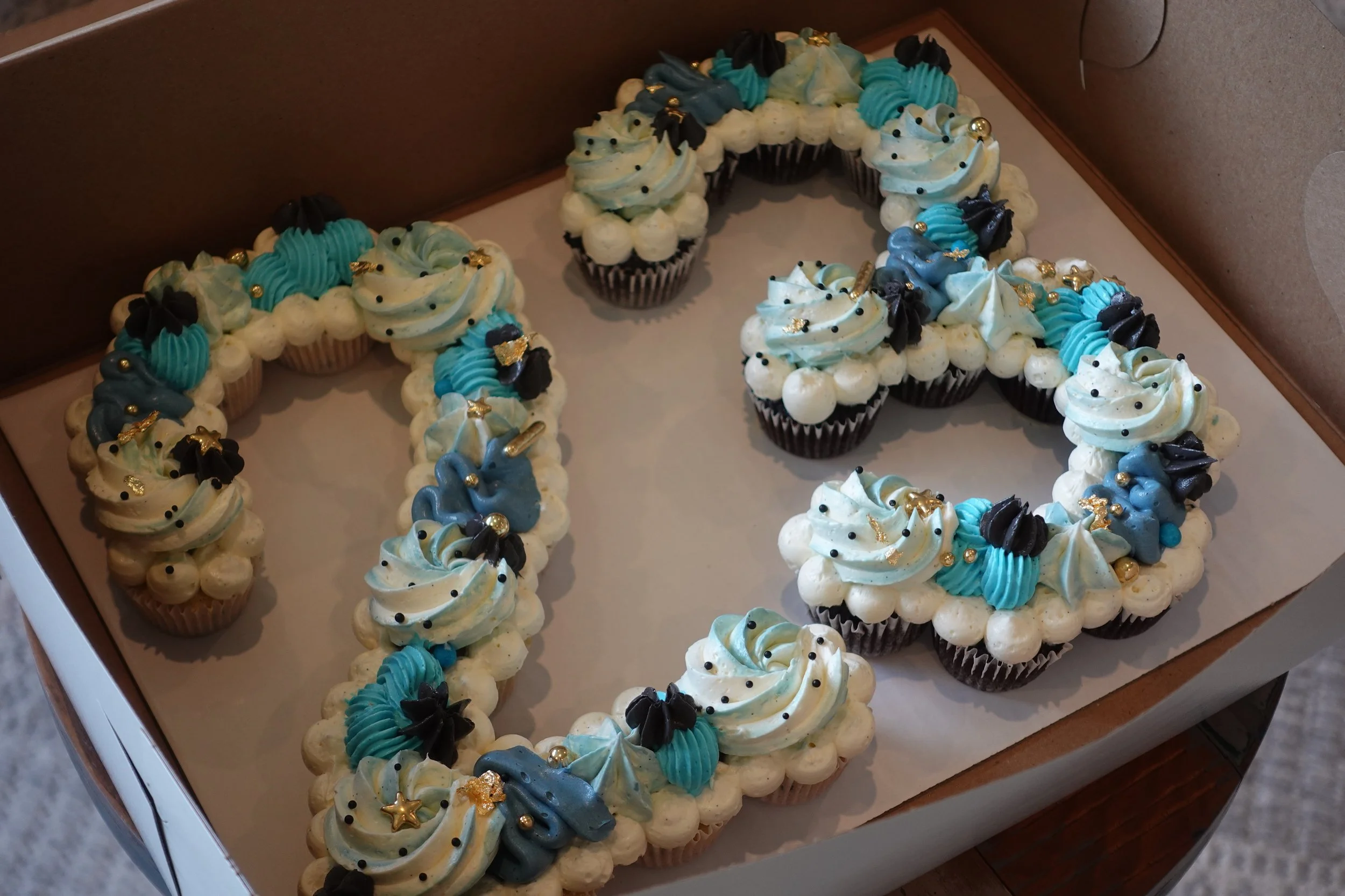 Cupcakes decorated with blue, black, white, and gold frosting and sprinkles, arranged in the shape of the number 4.