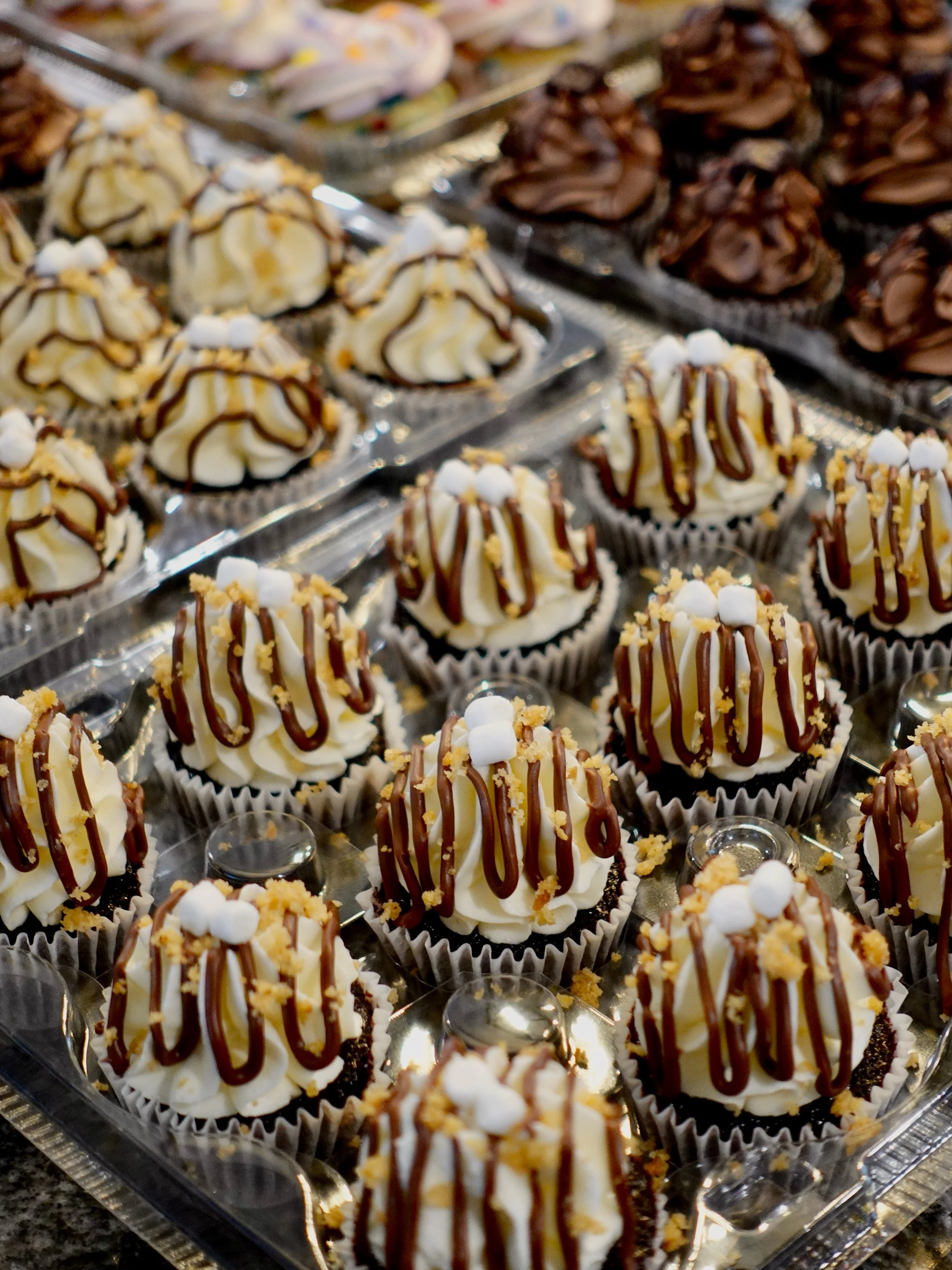 A tray of decorated cupcakes topped with white frosting, chocolate drizzle, small marshmallows, and crumbled cookies.