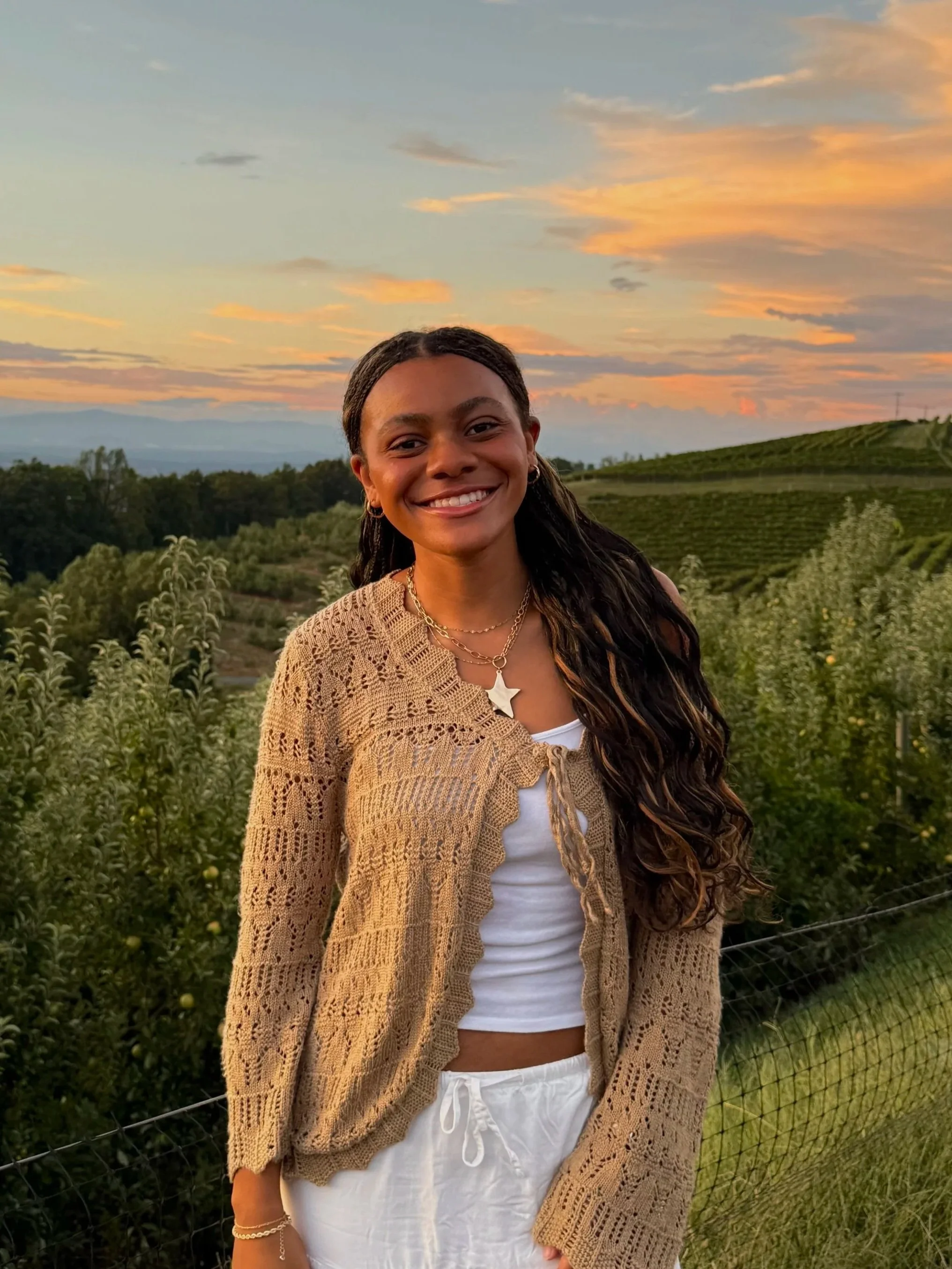 A young woman smiling outdoors during sunset, standing in front of a lush green landscape with a wire fence and rolling hills in the background.