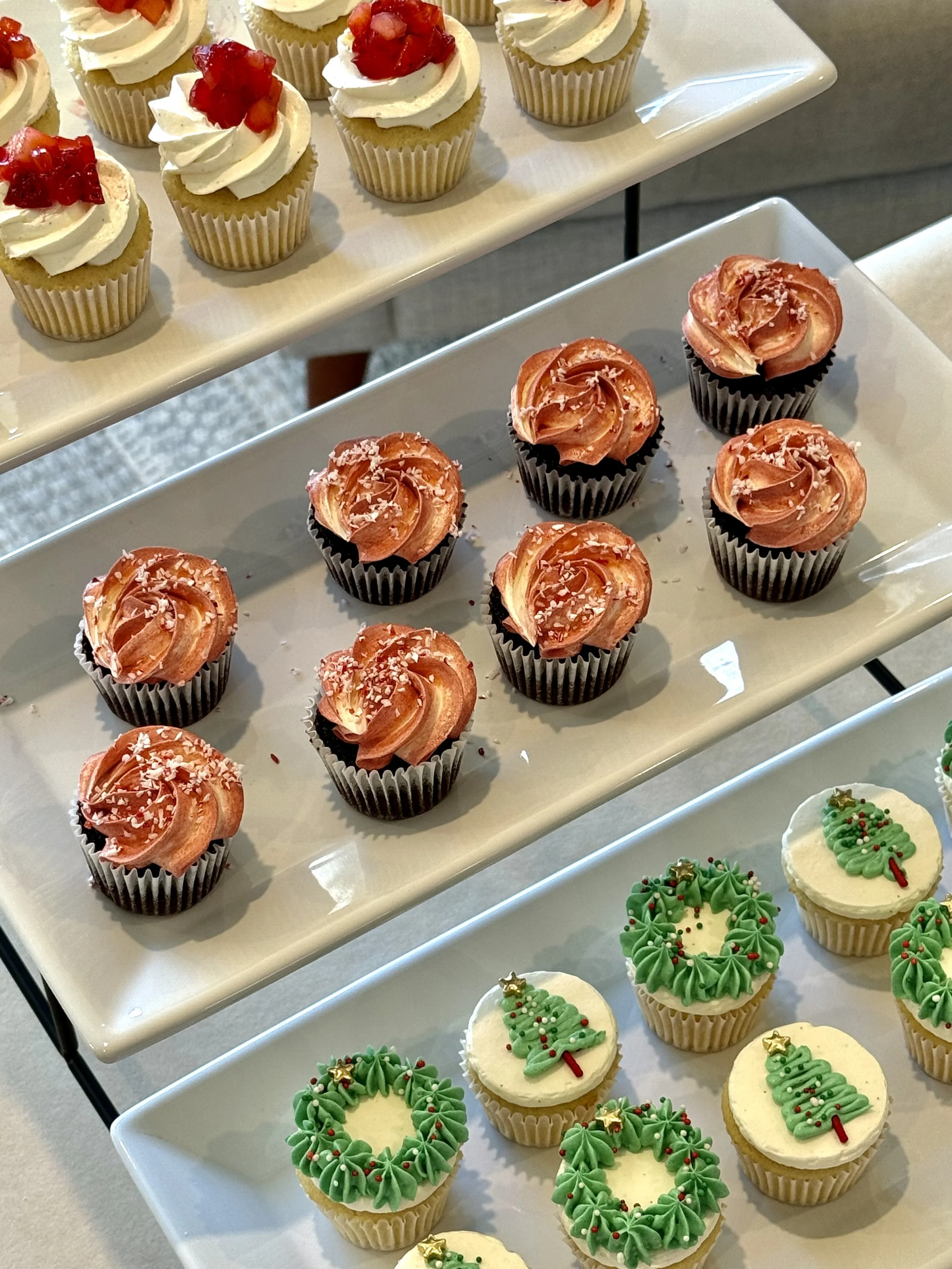 Assorted cupcakes on display, decorated with frosting, sprinkles, and holiday-themed toppings such as candy canes, Christmas trees, and wreaths.