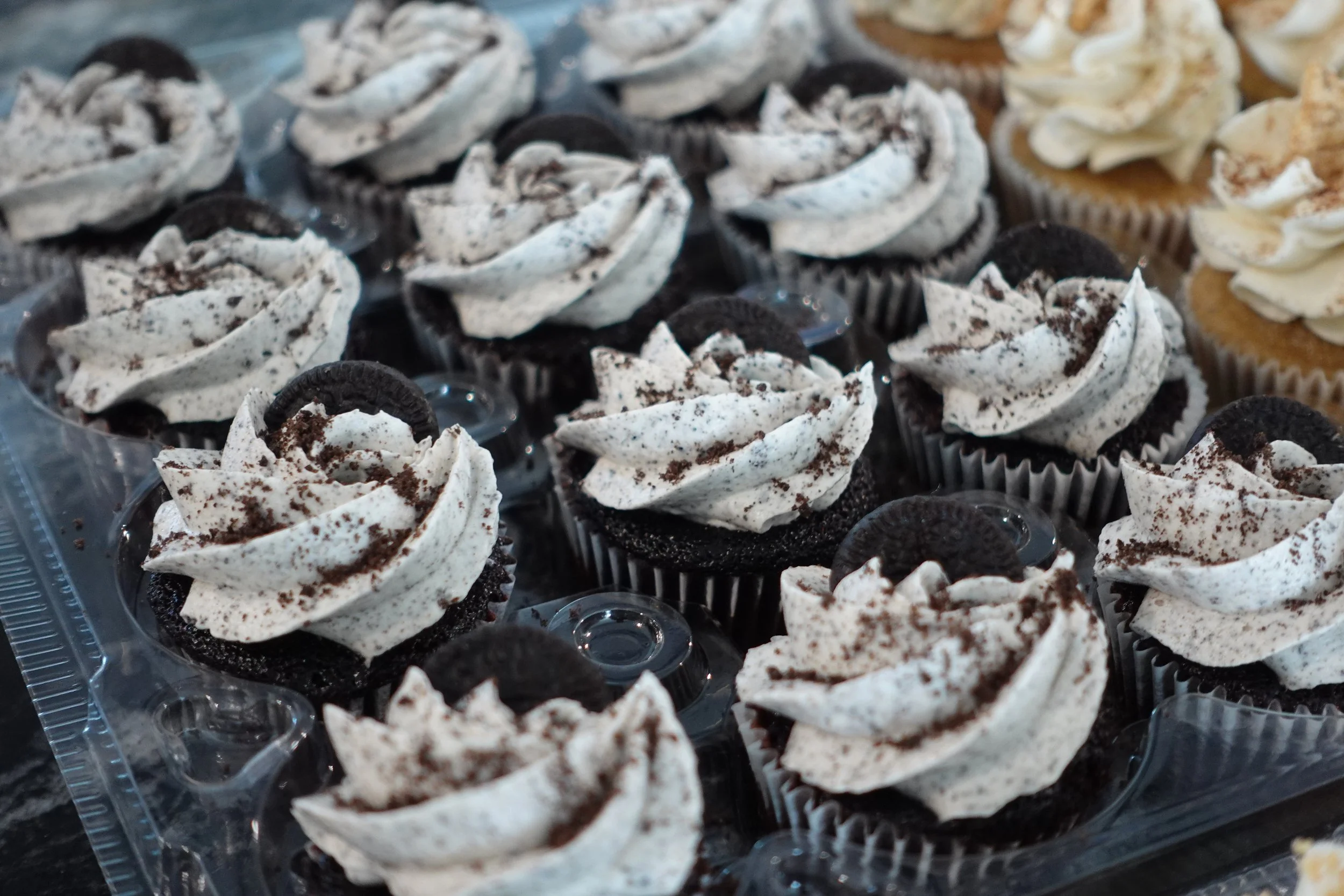Close-up of cupcakes with Oreo and vanilla frosting topped with crushed Oreo cookies in a plastic container.