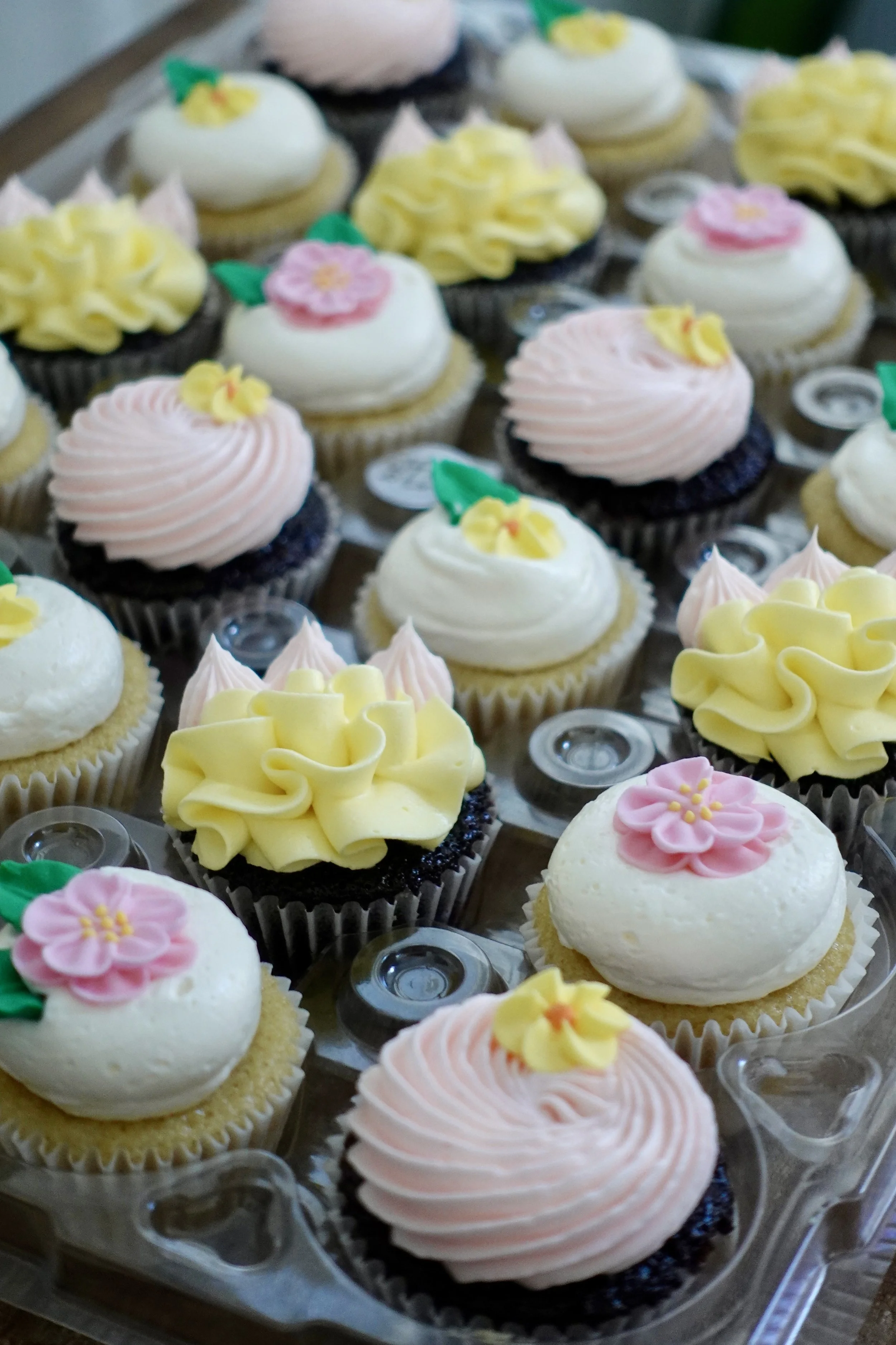 Assorted decorated cupcakes with pink, yellow, and white frosting, some topped with pink flowers and green leaves, arranged on a clear plastic tray.