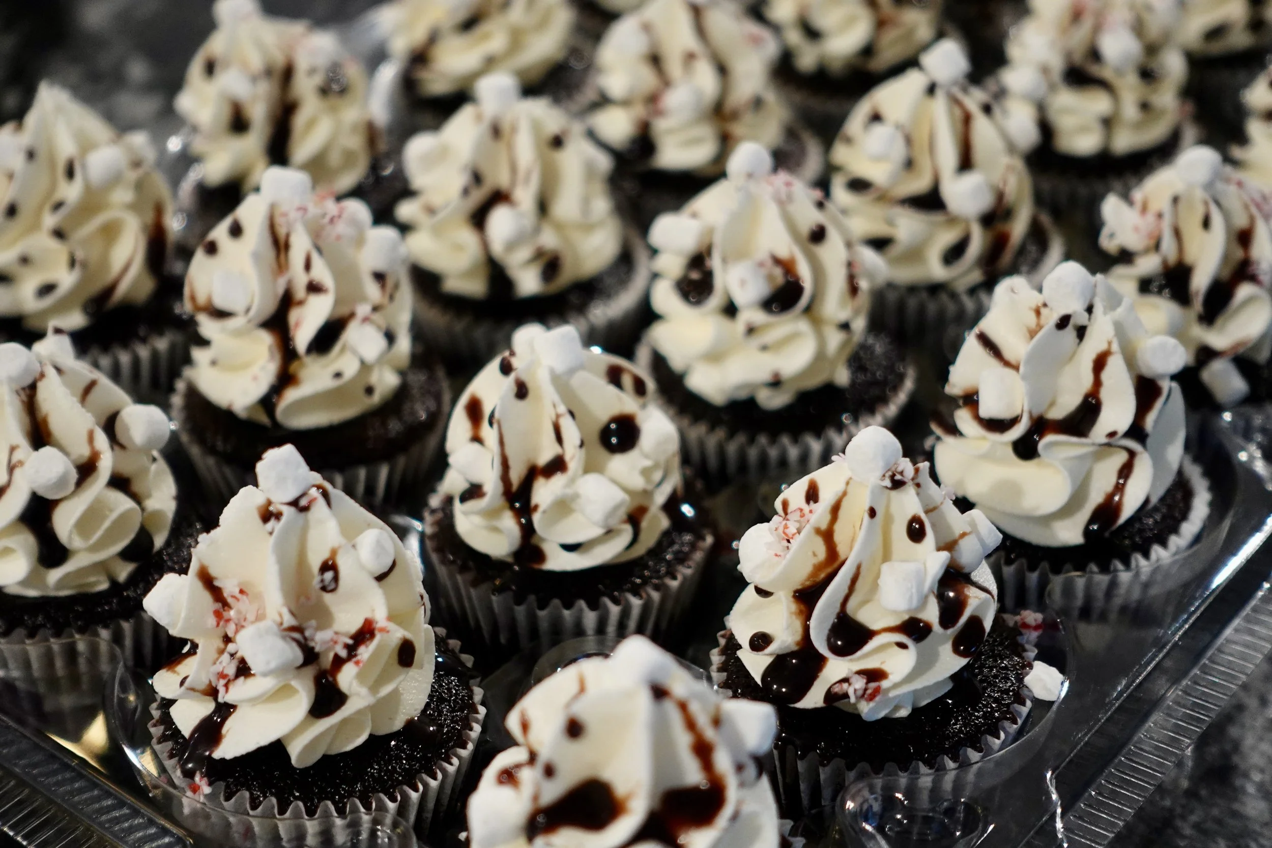 Assorted cupcakes with white frosting, chocolate drizzle, mini marshmallows, and red and white sprinkles on a tray.